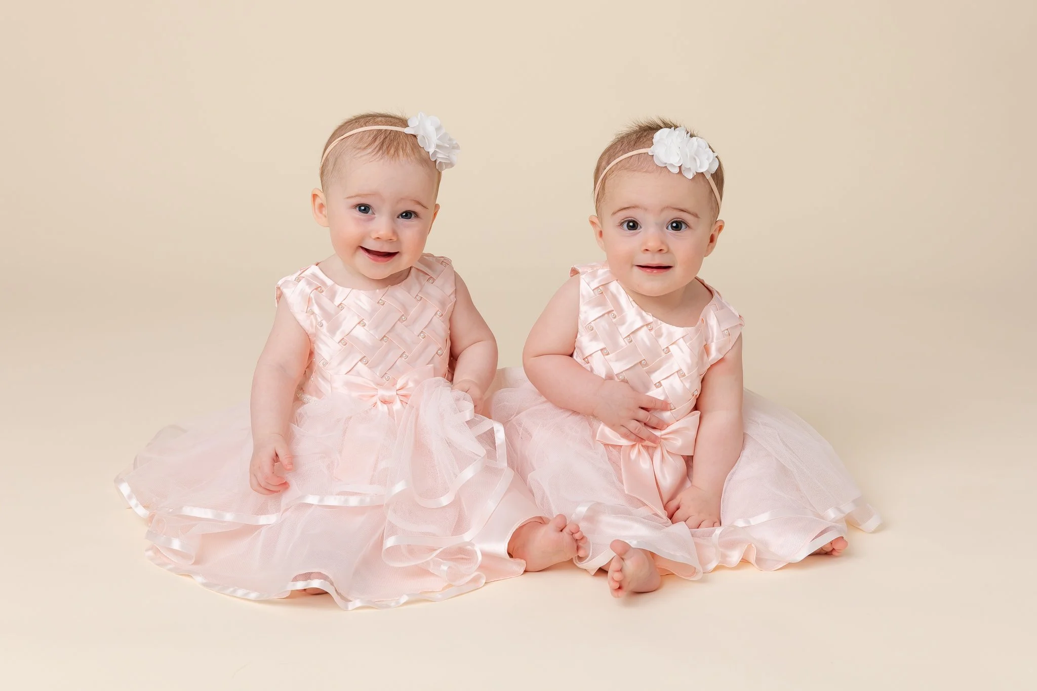 Two babies with light skin, wearing matching pink dresses and white flower headbands, sitting on a beige background.