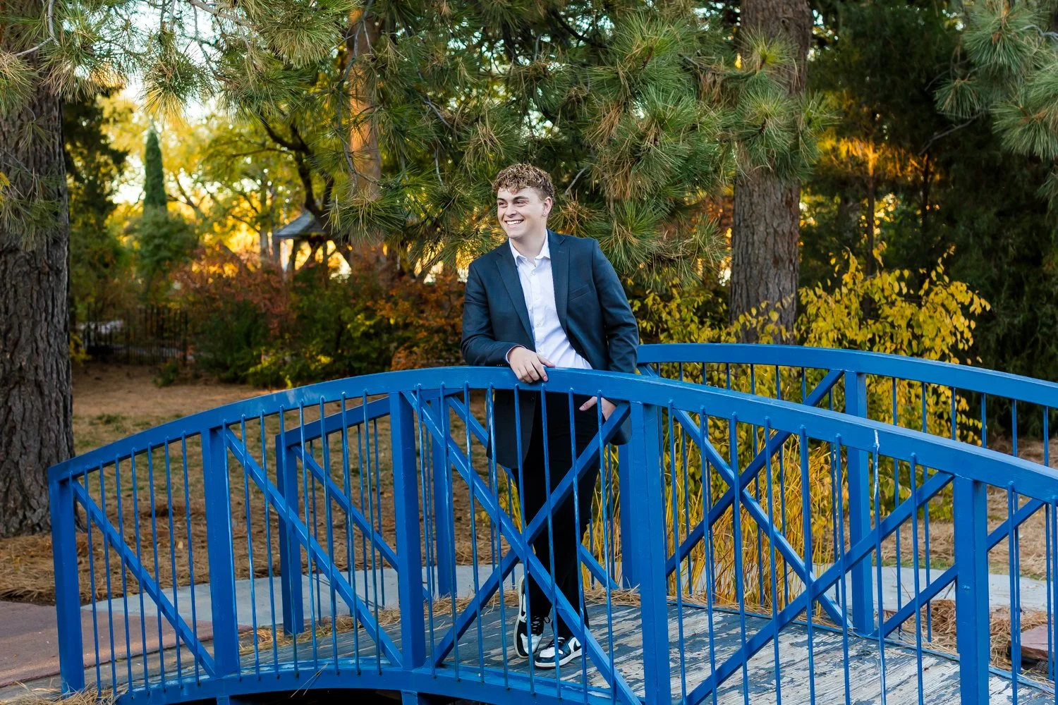 A young man in a suit leaning on a blue small arched bridge in a park with trees and fall foliage in the background.