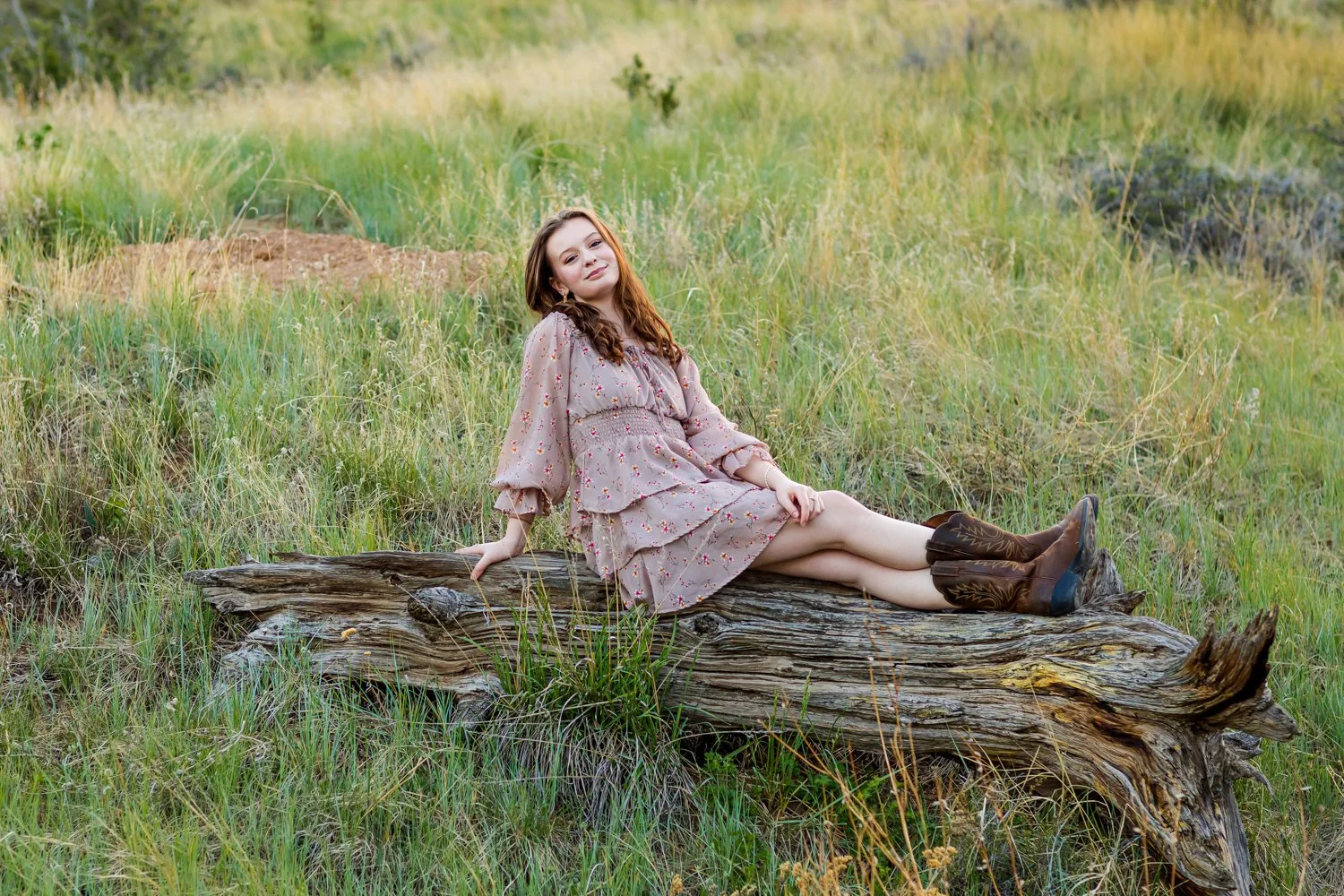 A young woman in a floral dress and cowboy boots sitting on a large fallen log in a grassy field.