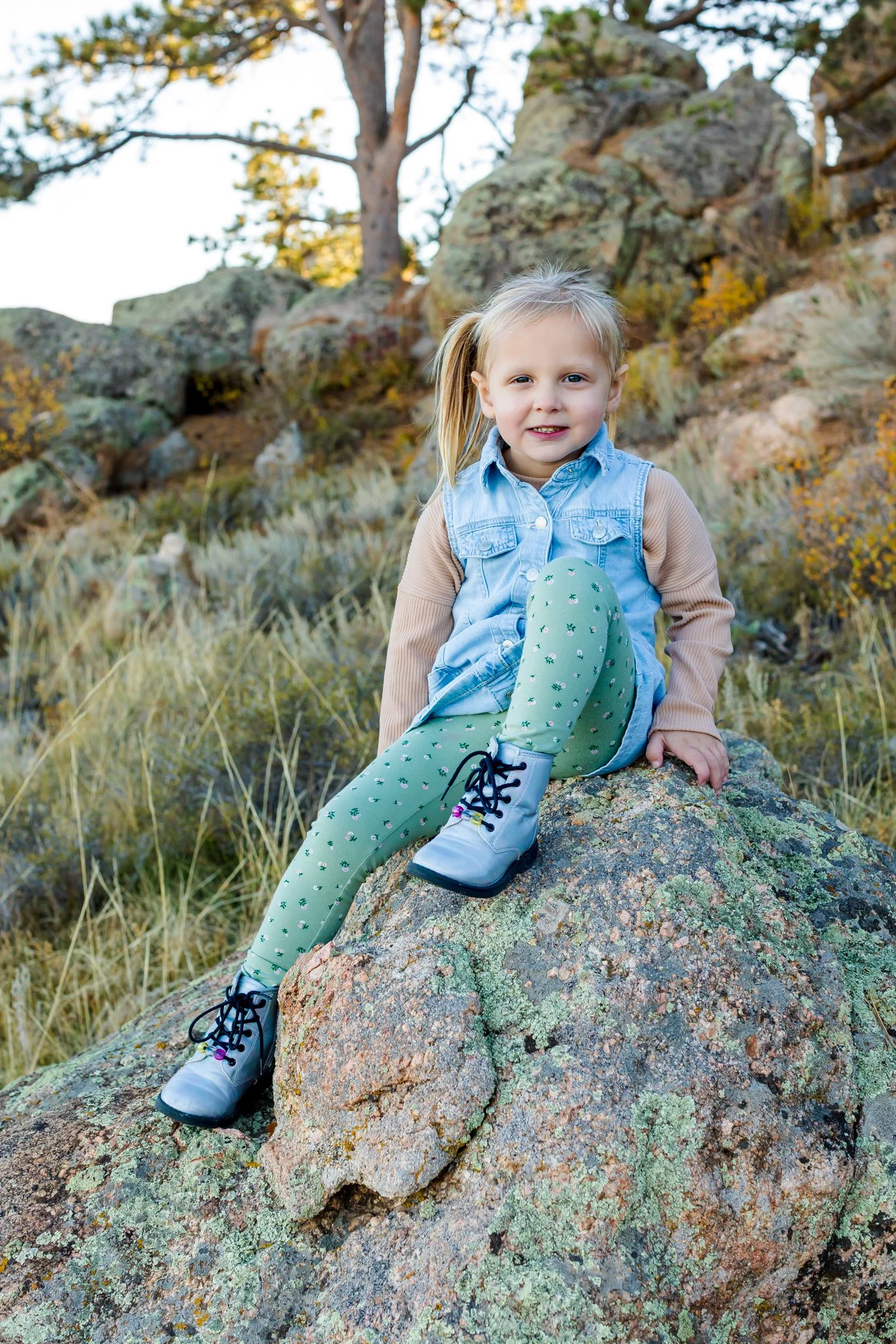 Young girl sitting on a large rock outdoors among trees and some autumn foliage, wearing a denim jacket, patterned leggings, and boots at Curt Gowdy Wyoming