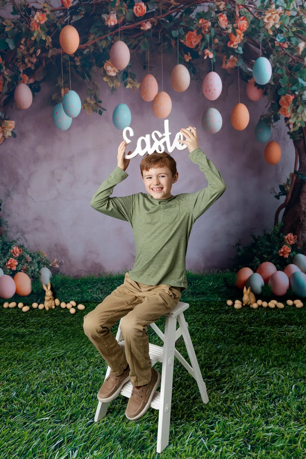 A smiling boy sitting on a white step ladder holding a white 'Easter' sign with hanging pastel-colored eggs and rabbit figures on a decorated outdoor background.