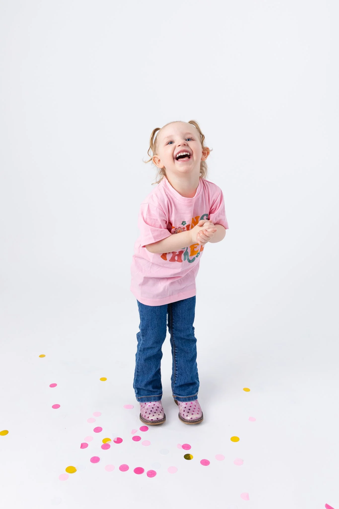 A young girl with light skin, wearing a pink T-shirt, jeans, and pink shoes with stars, is laughing and standing amidst scattered pink, yellow, and white confetti against a plain white background.