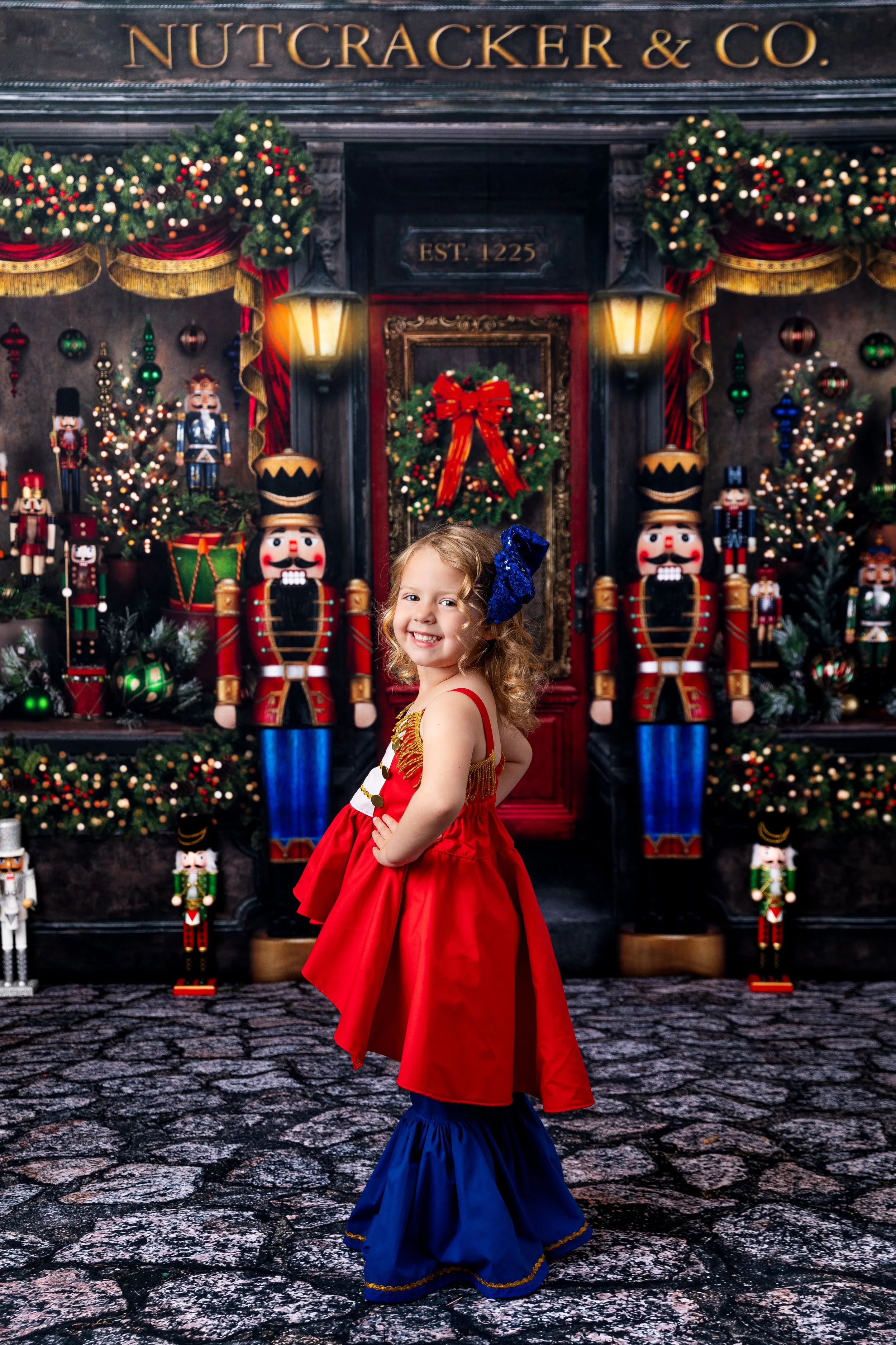 Young girl in a red and blue holiday dress standing in front of Nutcracker decorations and Christmas greenery with ornaments and lights, Christmas wreath, and Nutcracker figures at Nutcracker & Co. store in Cheyenne Studio.