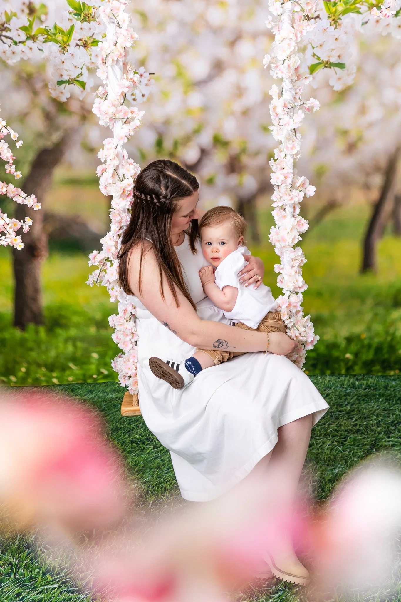 A woman with long brown hair holding a young child on her lap on a swing decorated with pink and white flowers, set against a background of blooming trees and green grass.
