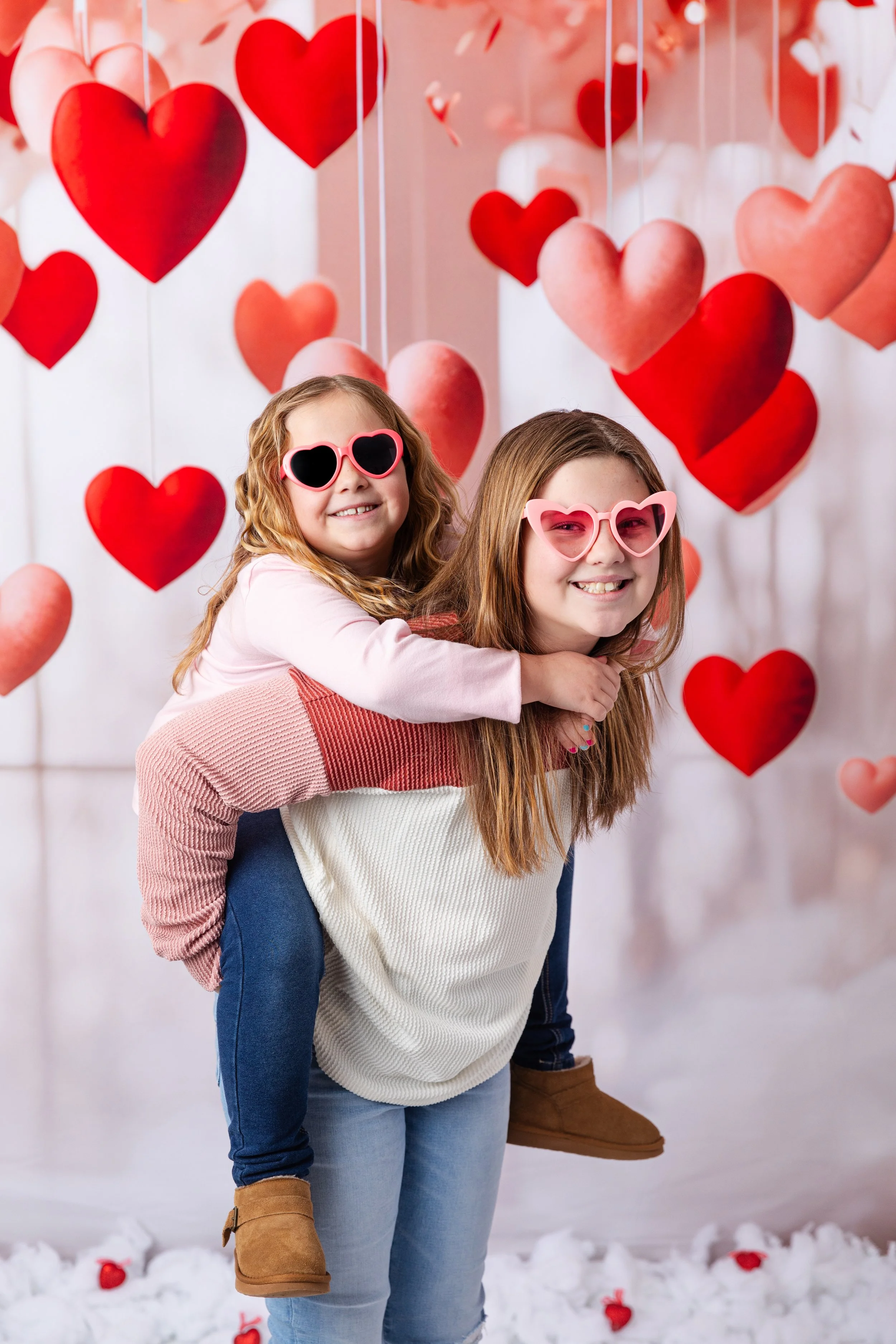 Two girls wearing pink heart-shaped sunglasses, smiling, with one giving the other a piggyback ride, surrounded by hanging red and pink heart decorations for Valentine's Day.