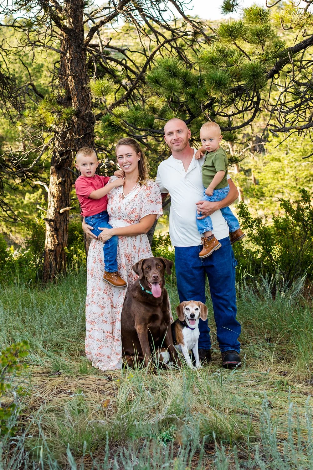 A family of four with two dogs outdoors in a wooded area, with trees and greenery in the background.