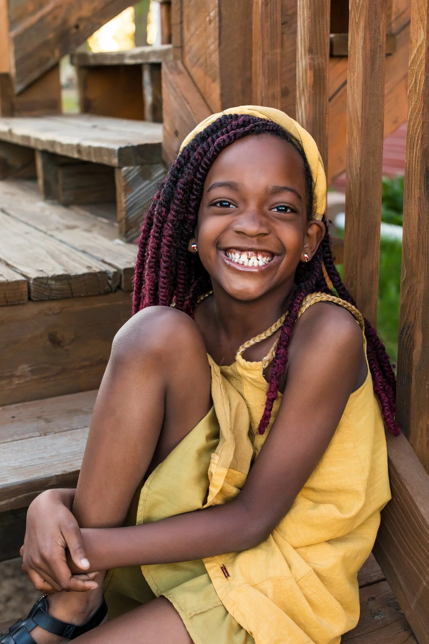 A young girl with dark braided hair, earrings, and a bright smile sitting on wooden stairs outdoors.
