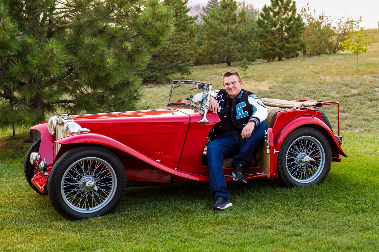 A smiling young man sitting on the door of a vintage red convertible car parked on a grassy field with pine trees in the background.