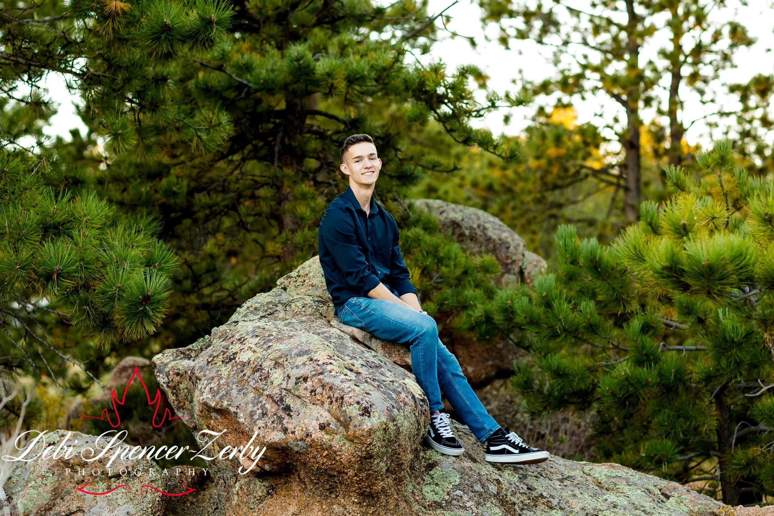 High School Senior sitting on a large rock in a forest with green pine trees, smiling, wearing a navy blue shirt and jeans at Curt Gowdy State Park, Wyoming