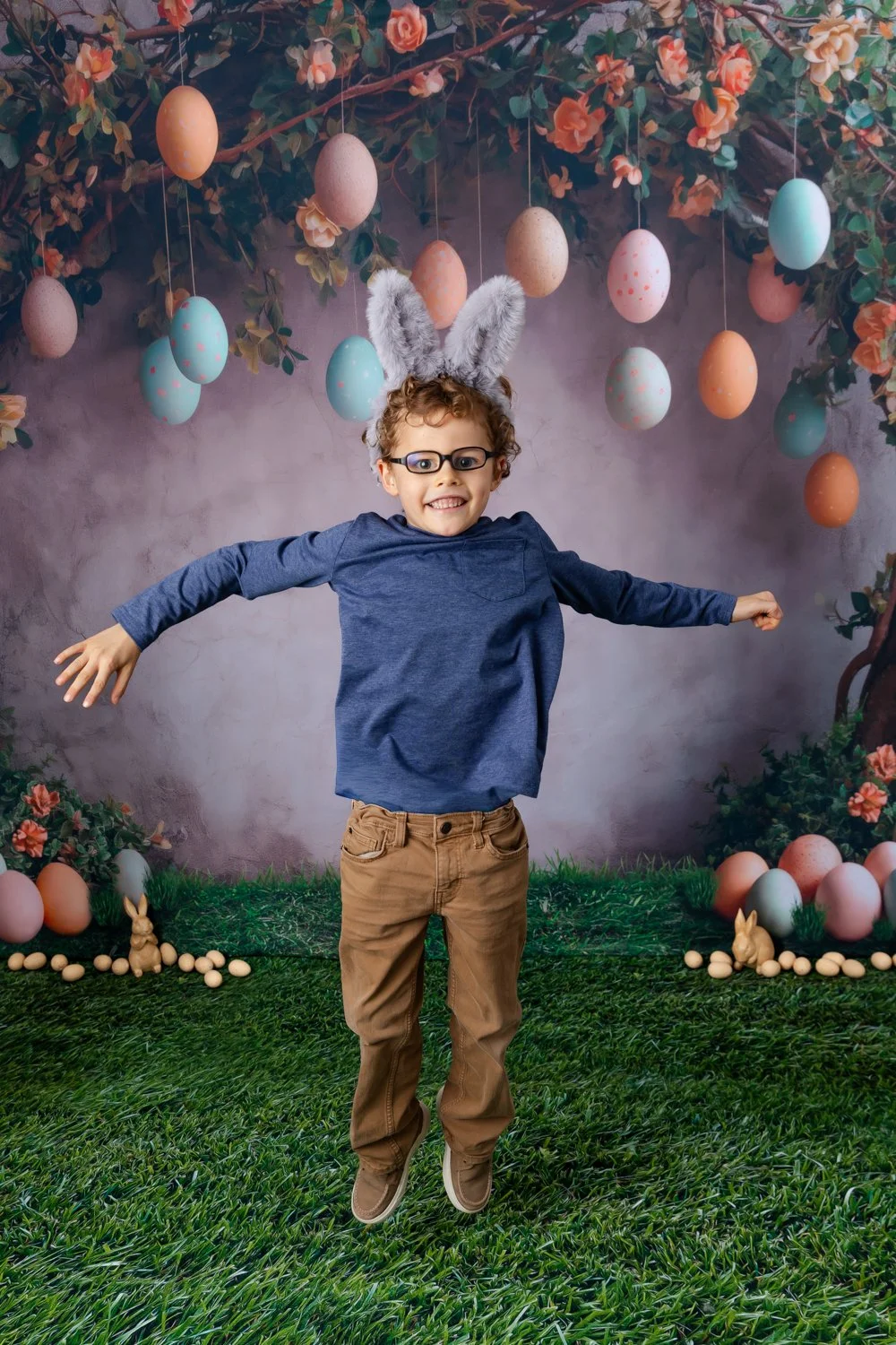 Child wearing glasses and bunny ears costume jumping with arms outstretched in front of Easter-themed backdrop with decorated eggs and bunnies, grass floor.