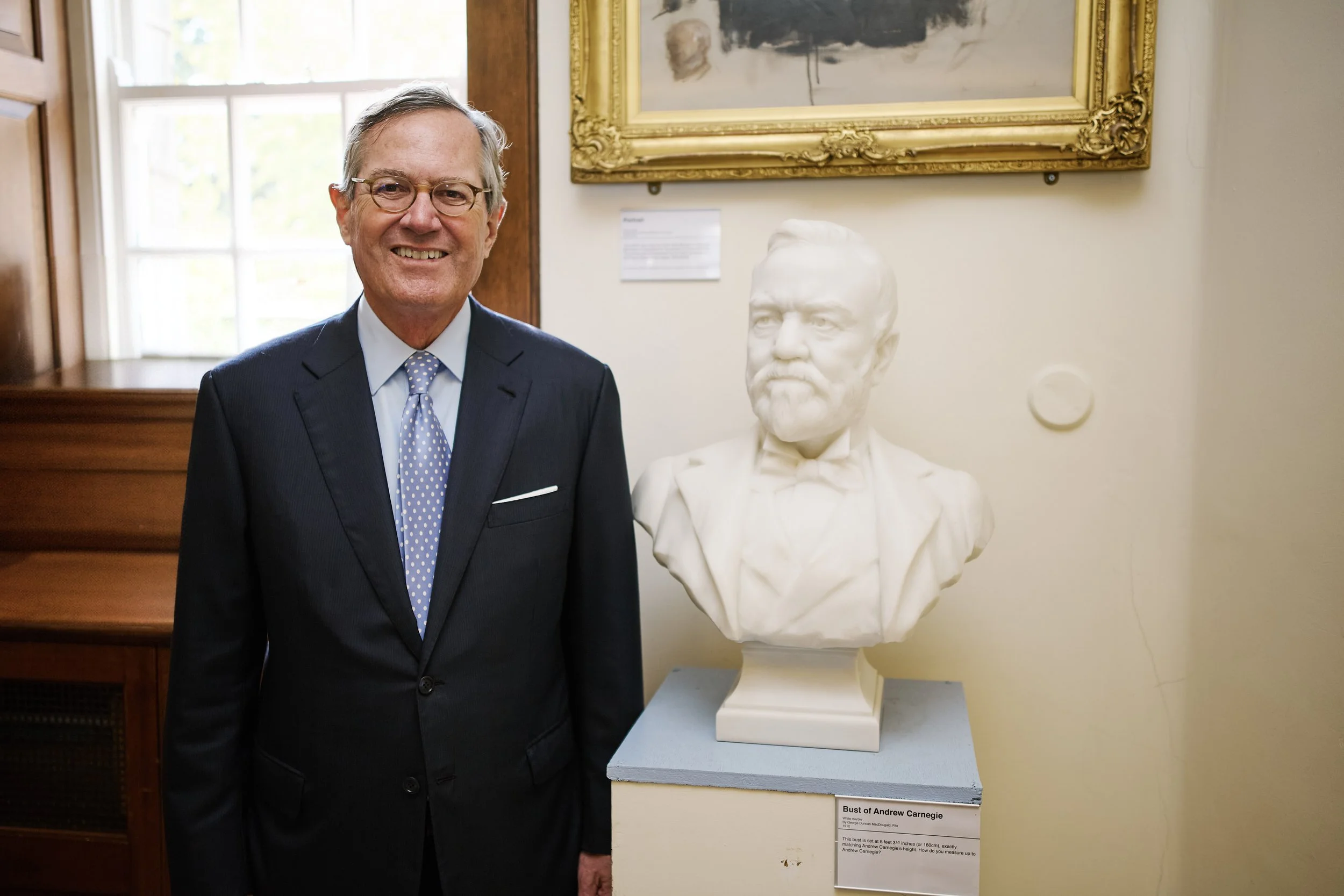 White man in 60s wearing a navy blue suit, light blue shirt and tie stands beside a white marble bust of Andrew Carnegie, crafted for when Carnegie was in his 60s