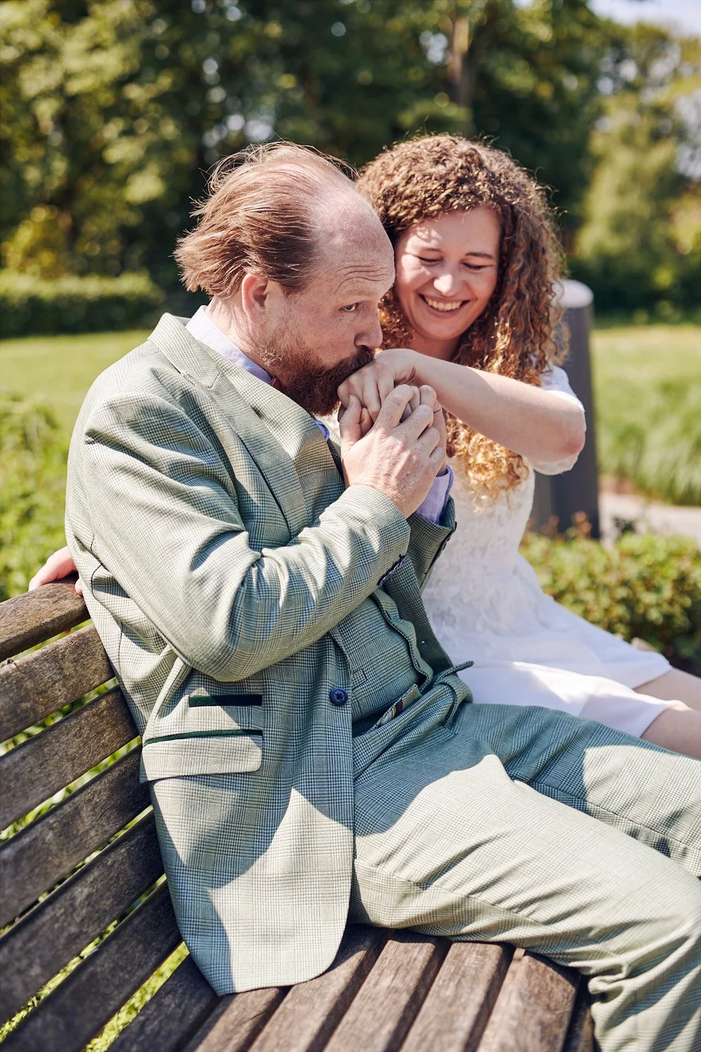 Groom kissing brides hand while sitting on a park bench.