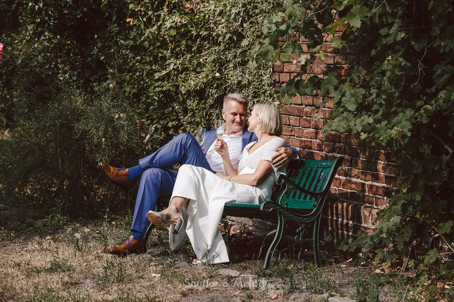 A mature bride and groom sitting on a bench toasting each other.