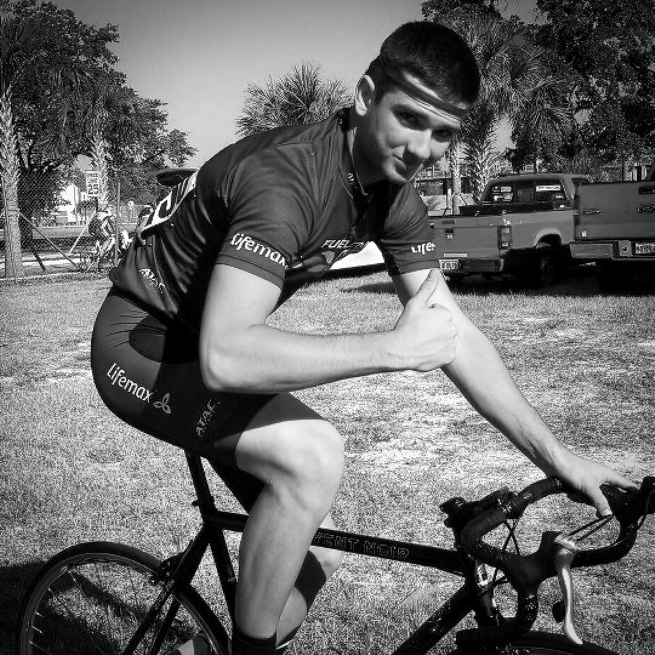 A young cyclist with short dark hair and a GUTR performance sweatband leaning on a bicycle handlebar, outdoors with trees and parked cars in the background, smiling, giving a thumbs up with his right hand.
