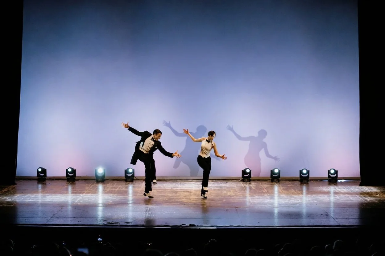 Due persone che ballano in un palco con luci sullo sfondo, indossano abiti eleganti e sono in movimento orchestrato.