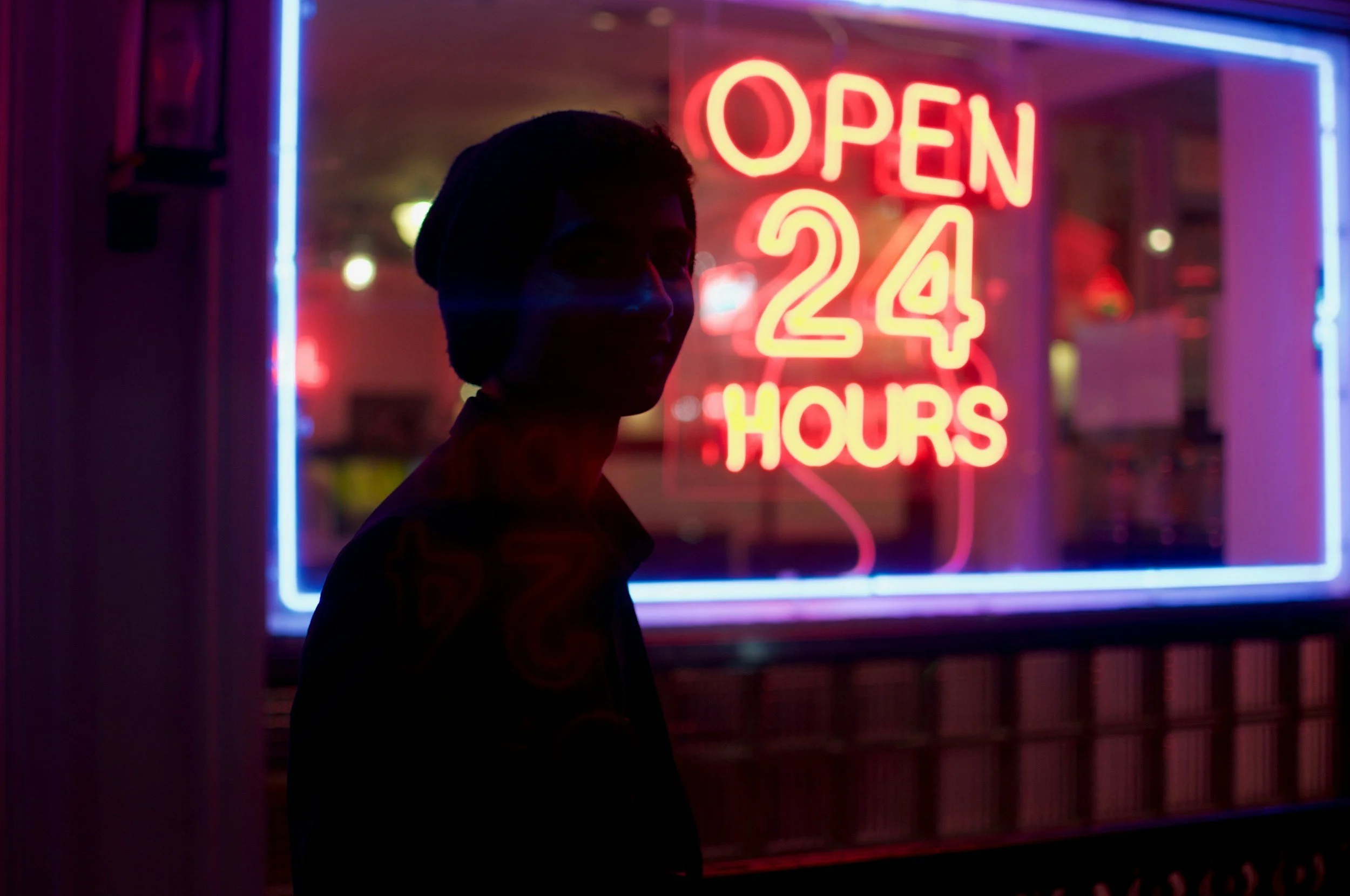 Silhouette of a person standing outside a storefront with a bright red neon sign indicating "Open 24 Hours".