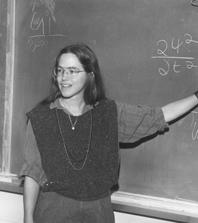 A young woman (Blount) with glasses, shoulder-length hair, wearing a sweater vest over a shirt, standing in front of a blackboard with her arm extended, as she teaches or presents.