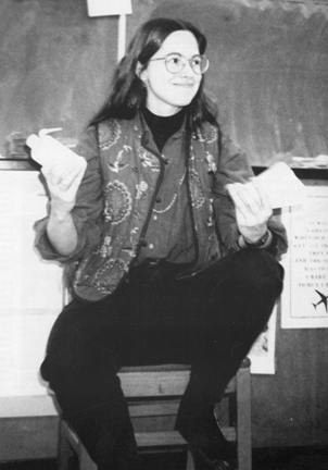 A woman (Blount) sitting on a chair in a classroom, holding a piece of paper in one hand and a water bottle in the other, wearing glasses and a vest.