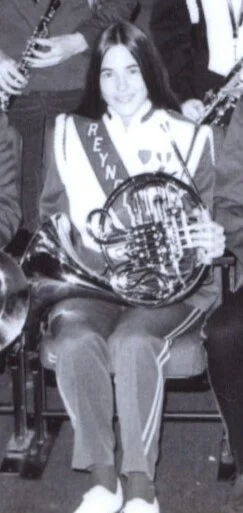 A young girl (Blount) sitting down holding a French horn, wearing a school band uniform.