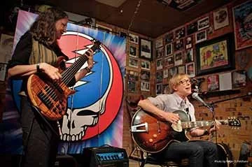 Two musicians performing: a woman with curly hair playing an electric bass and anothe with short hair and glasses singing and playing an acoustic guitar, in a room decorated with framed pictures and a large Grateful Dead skull logo in the background.
