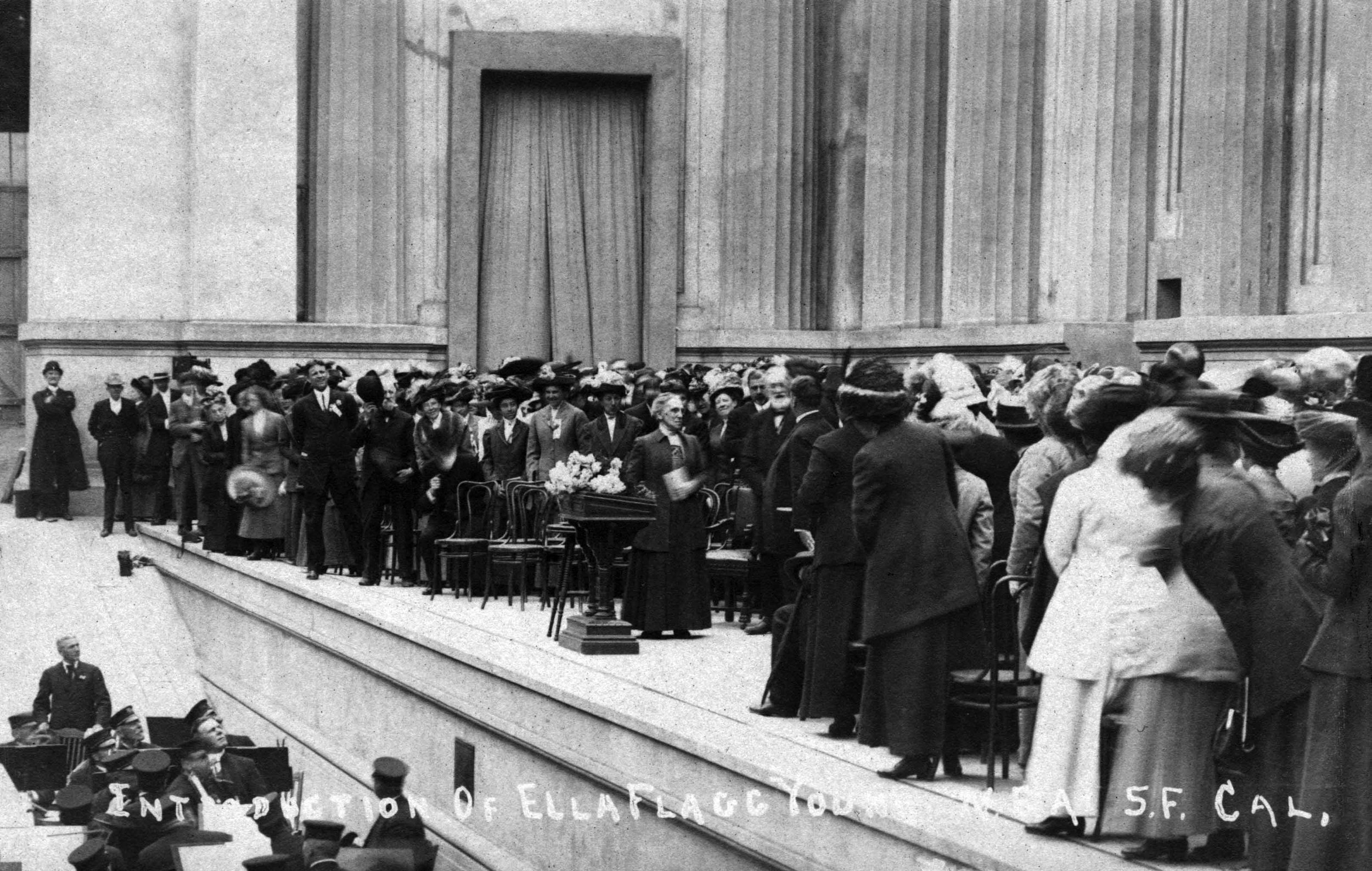 A historical black-and-white photograph showing a large gathering of women in early 20th-century attire, standing on an outdoor stage with a large building facade in the background. The scene appears to be a public demonstration or event, with some women holding flowers and others engaging with the crowd below.