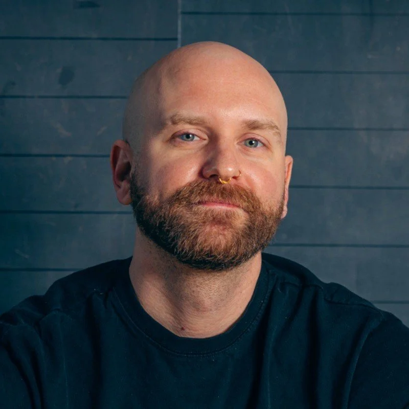 A bald man with a full beard and mustache, wearing a nose ring and necklace, in front of a blue wooden wall.
