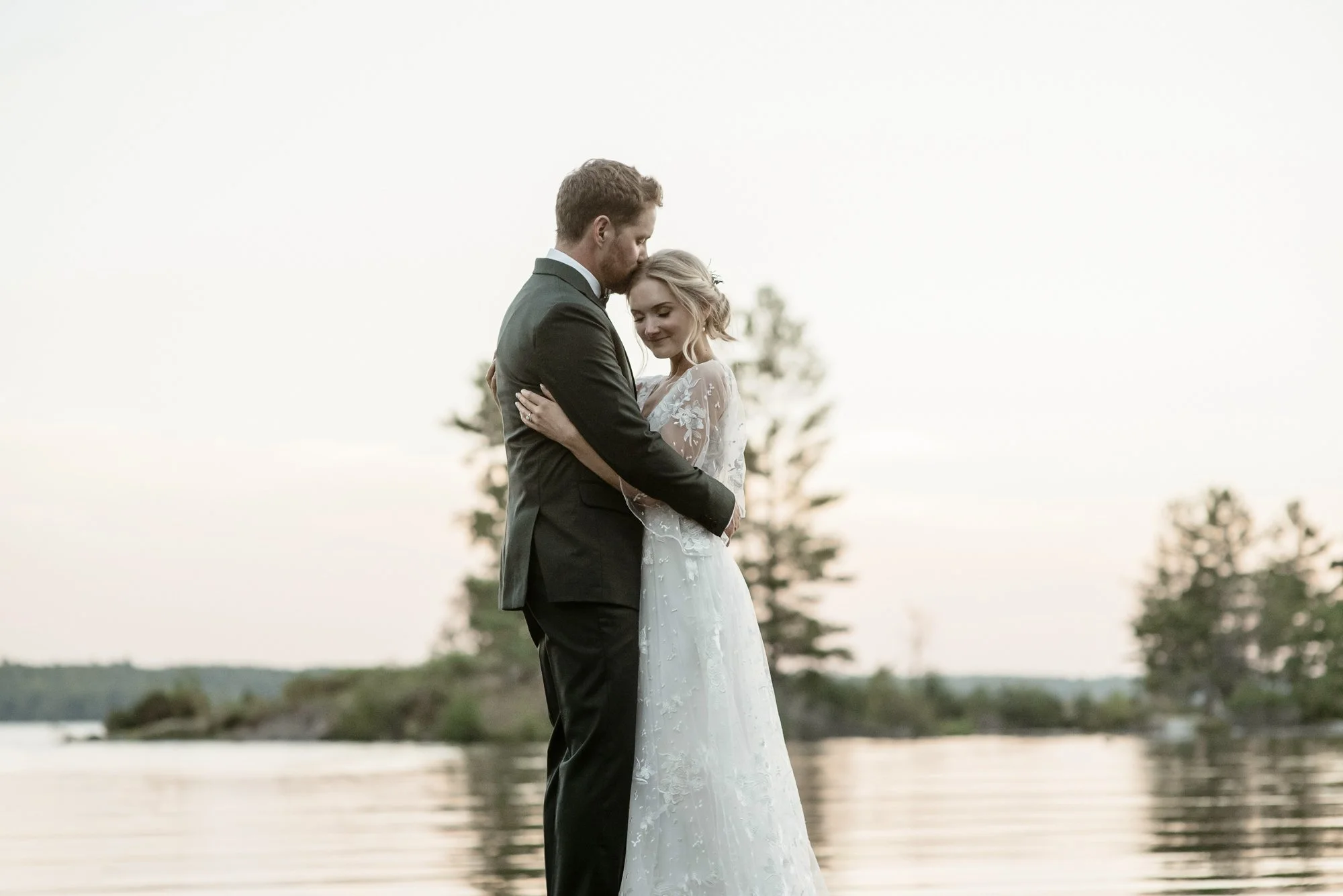 A bride and groom standing close together outdoors by a river, with the groom gently kissing the bride's forehead during sunset.