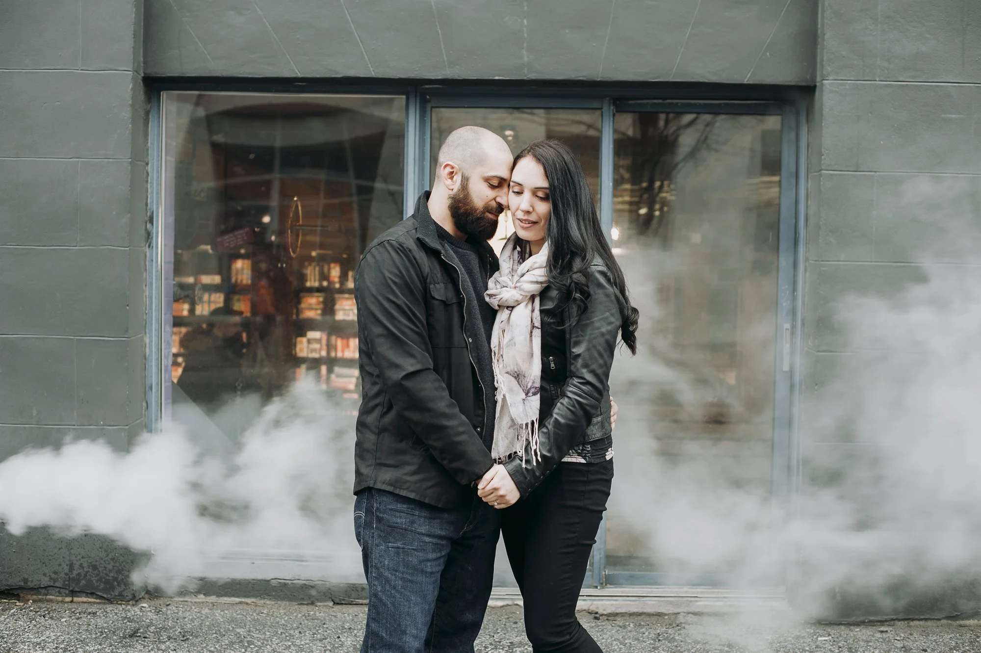 A man and a woman stand close together, holding hands with their foreheads touching in front of a building with a window. Steam is visible around their feet.