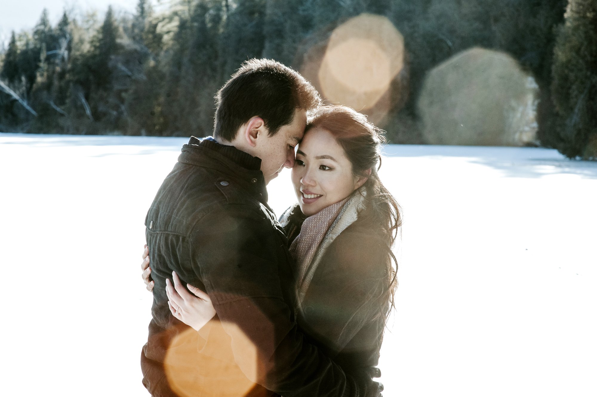 A couple embraces outdoors with a snowy landscape and forest in the background, sunlight creating lens flare effects.
