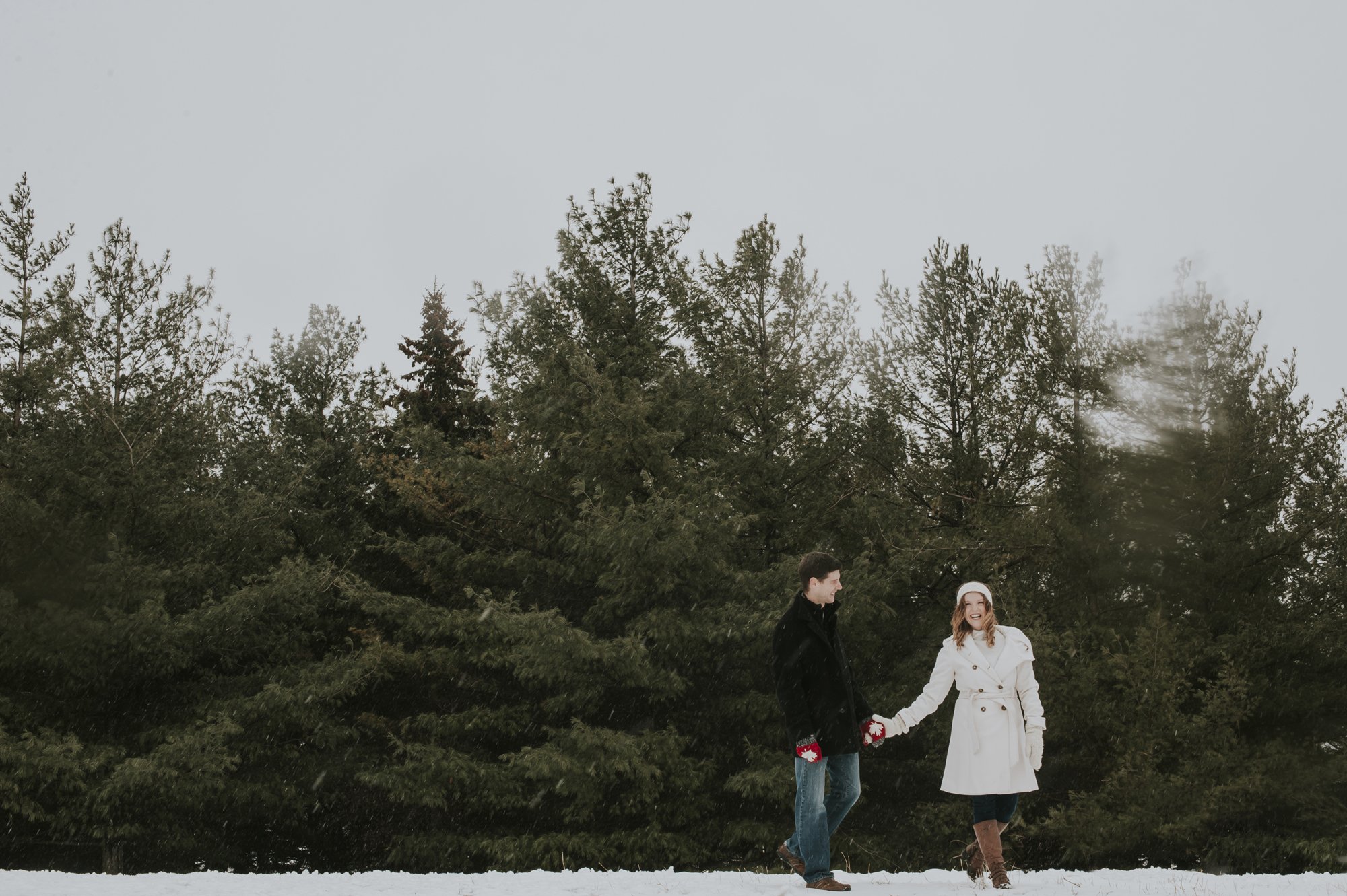 A couple holding hands outdoors in winter, standing on snow near tall pine trees, smiling, with the woman wearing a white coat and brown boots, and the man wearing a dark coat and jeans.