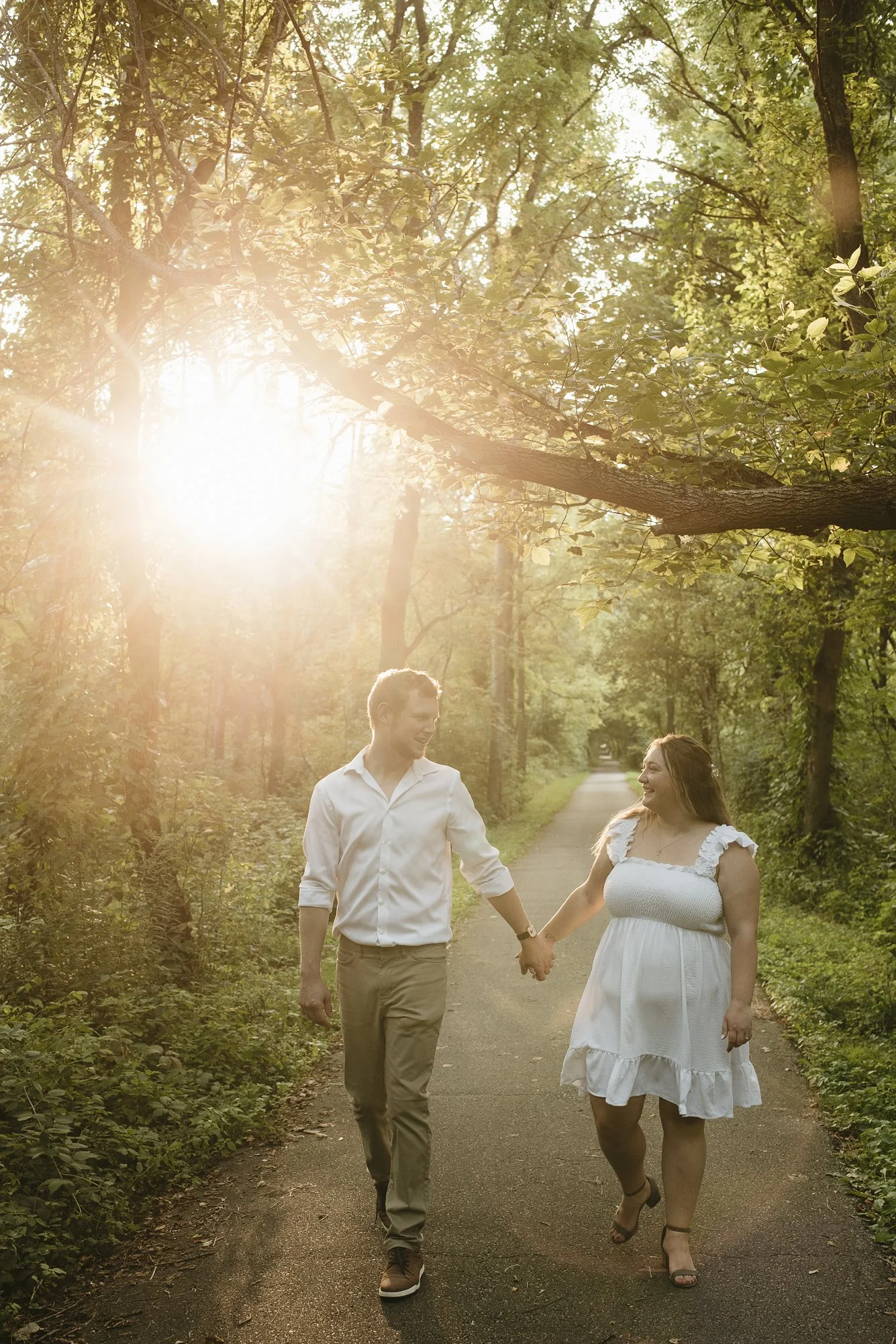 A couple walking hand in hand on a forest path during sunset, surrounded by green trees, with sunlight filtering through the leaves.