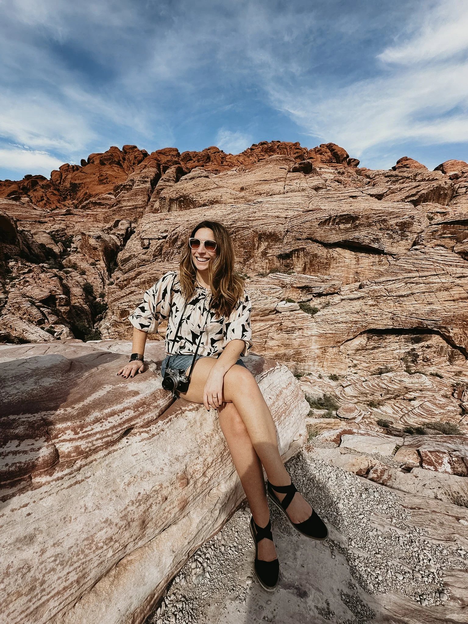 Woman sitting on a rock formation in a desert with red and beige layered rocks in the background, wearing sunglasses, a patterned shirt, shorts, and black shoes, smiling and holding a camera.