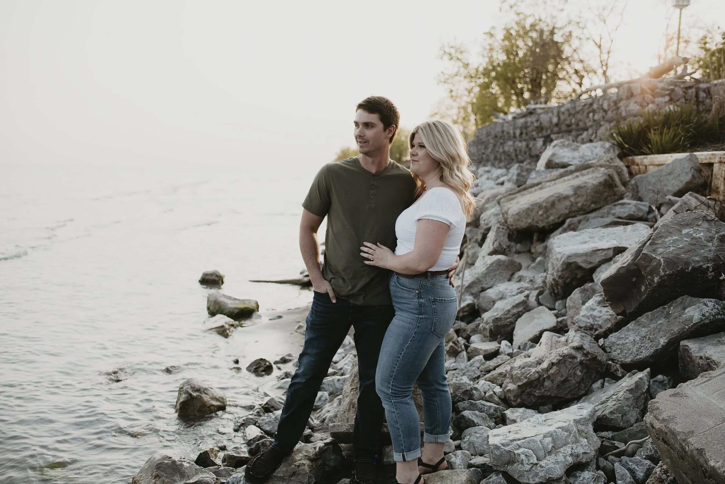 A young couple standing on rocks along a shoreline during sunset, looking at each other and smiling.