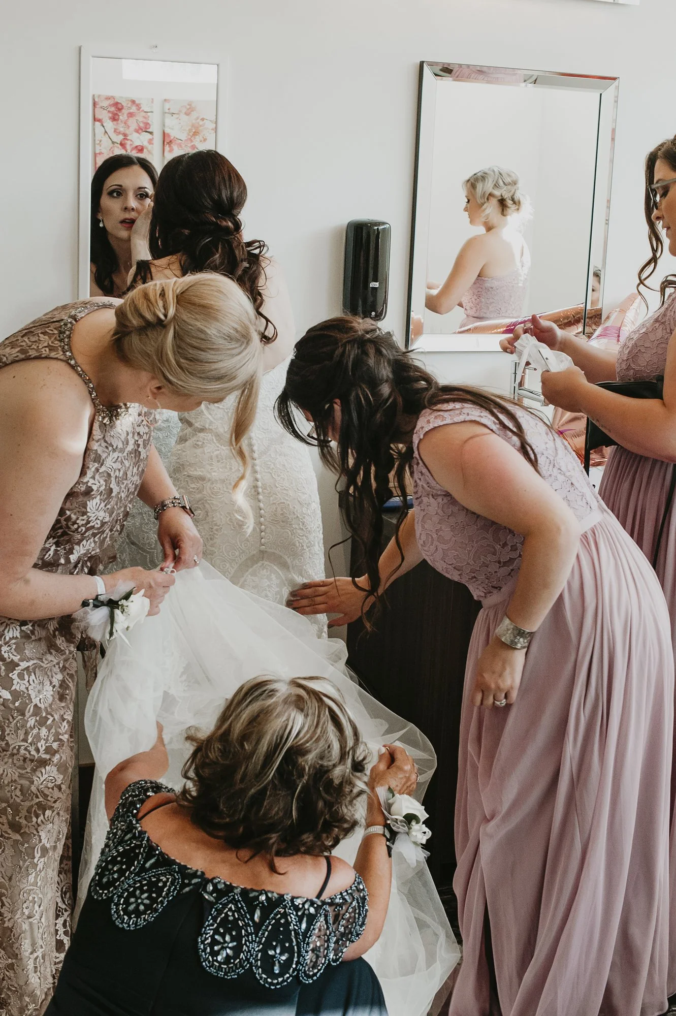 Group of women getting ready for a wedding, with some looking at the bride's dress while others prepare accessories, in a room with a mirror and wall art.