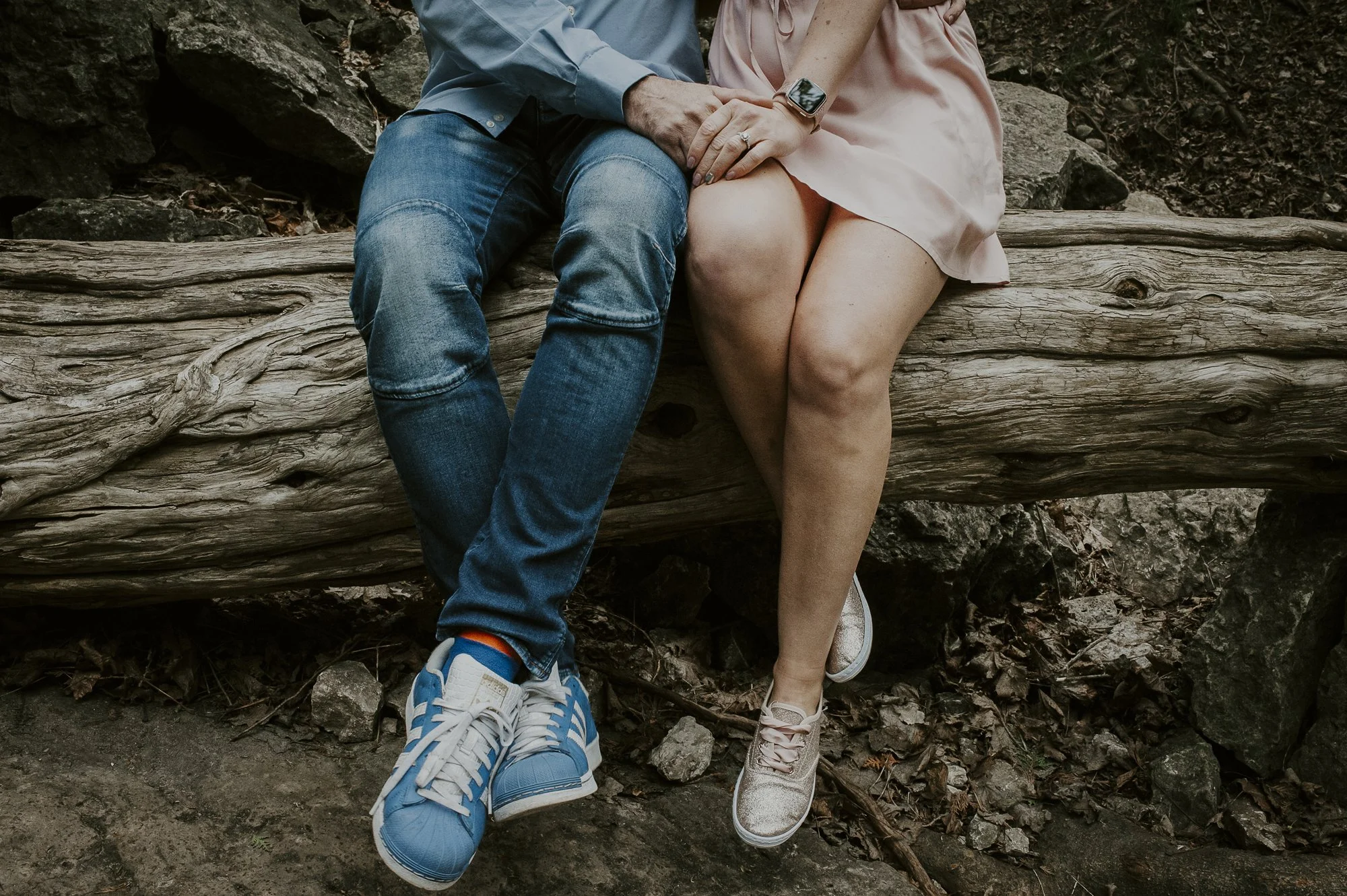 Close-up of a couple sitting on a fallen log outdoors. They are holding hands, with the woman wearing a smartwatch and a ring, and the man wearing blue sneakers and jeans. The woman is dressed in a pink dress, and the man in a blue shirt and jeans. T
