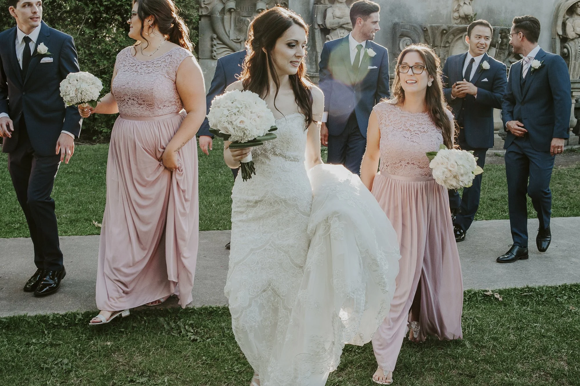 A group of people dressed in formal attire, including a bride in a white lace wedding dress holding a bouquet of white roses, bridesmaids in pink dresses also holding bouquets, and groomsmen in dark suits with white shirts and ties, walking outdoors 