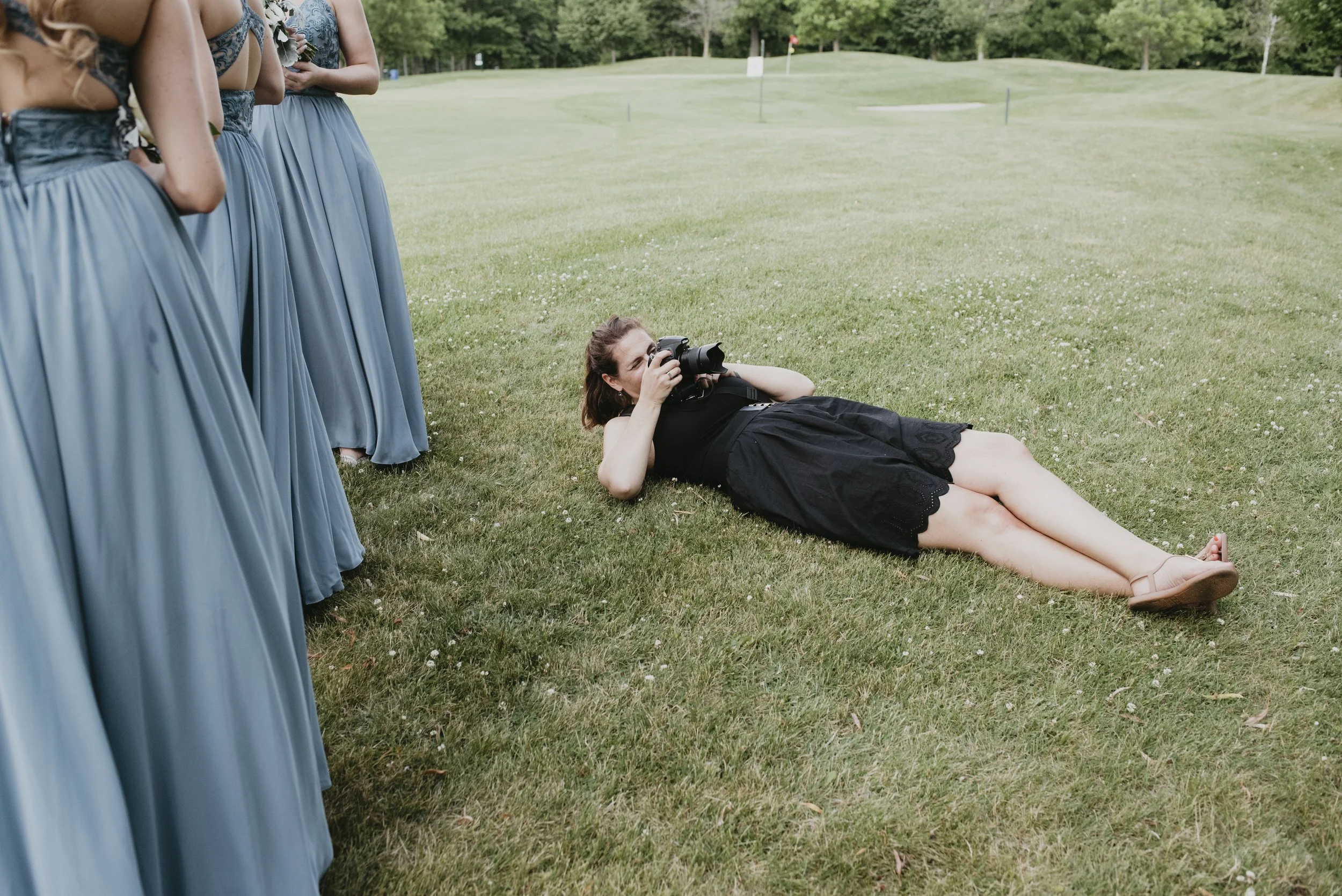A woman lying on the grass taking a photograph of four women wearing matching blue dresses at a golf course.
