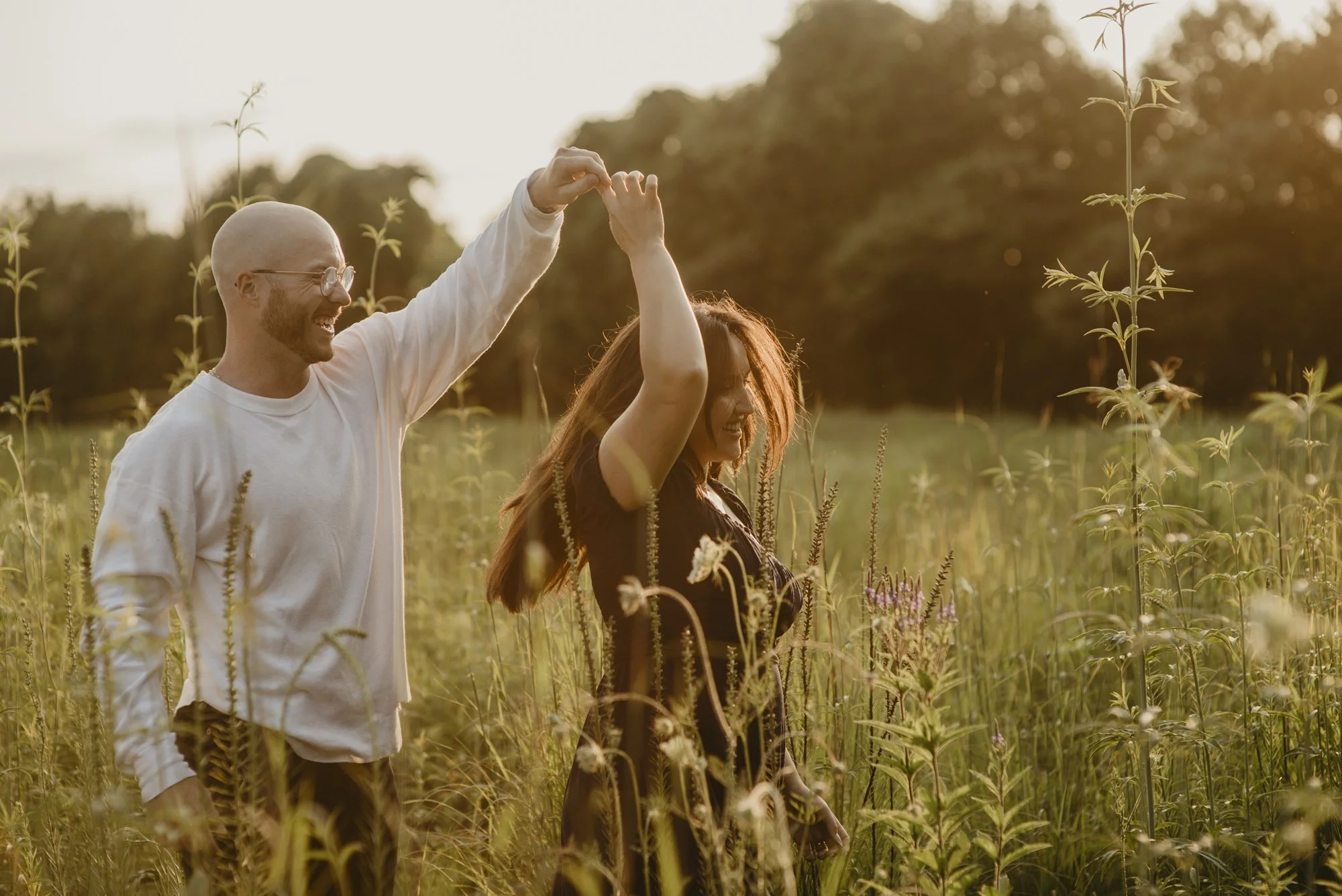 A man and woman holding hands and smiling in a field of tall plants during sunset.