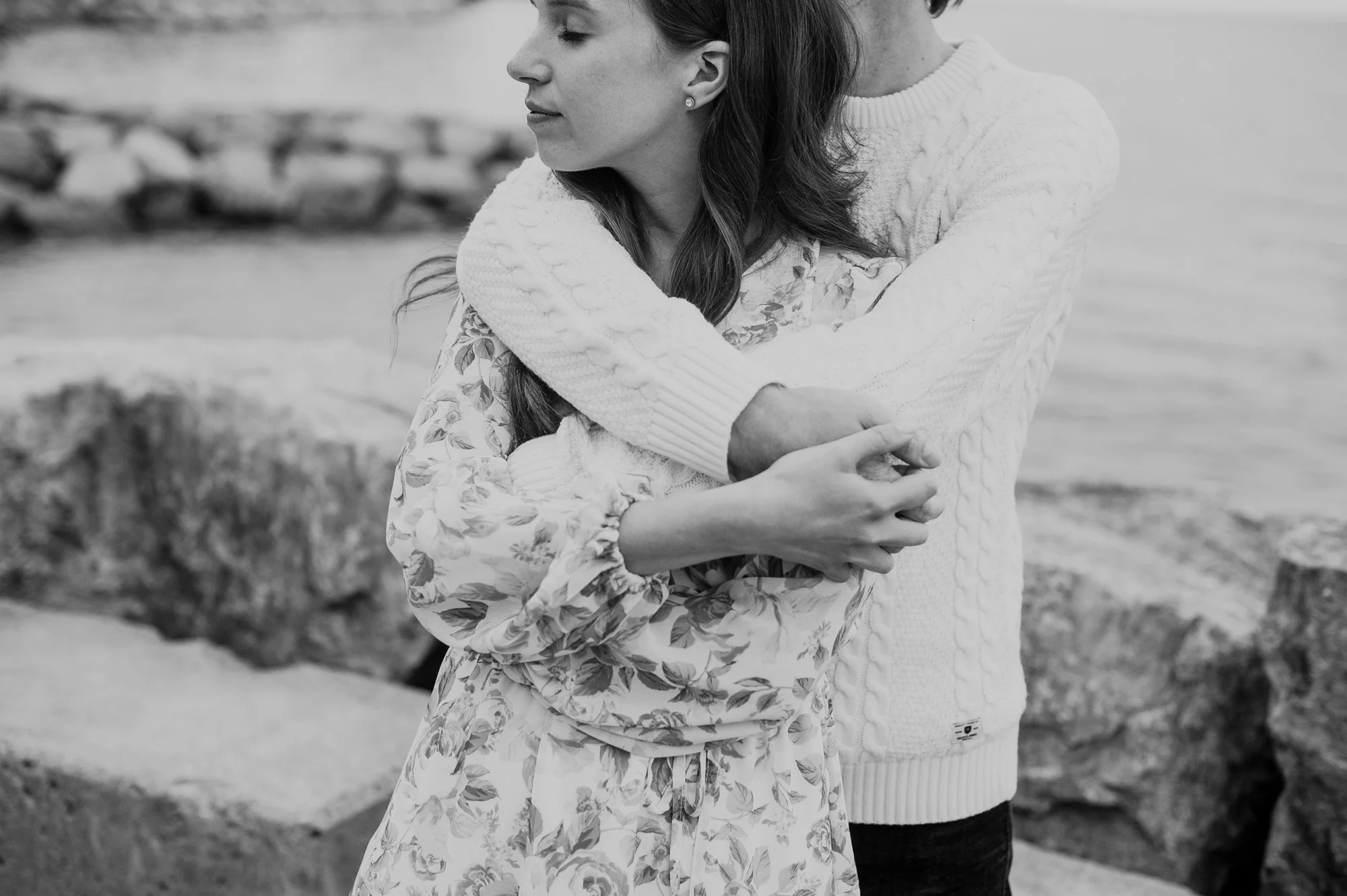 A black and white photo of a woman with long hair, wearing a floral dress, being embraced by a person in a white sweater.