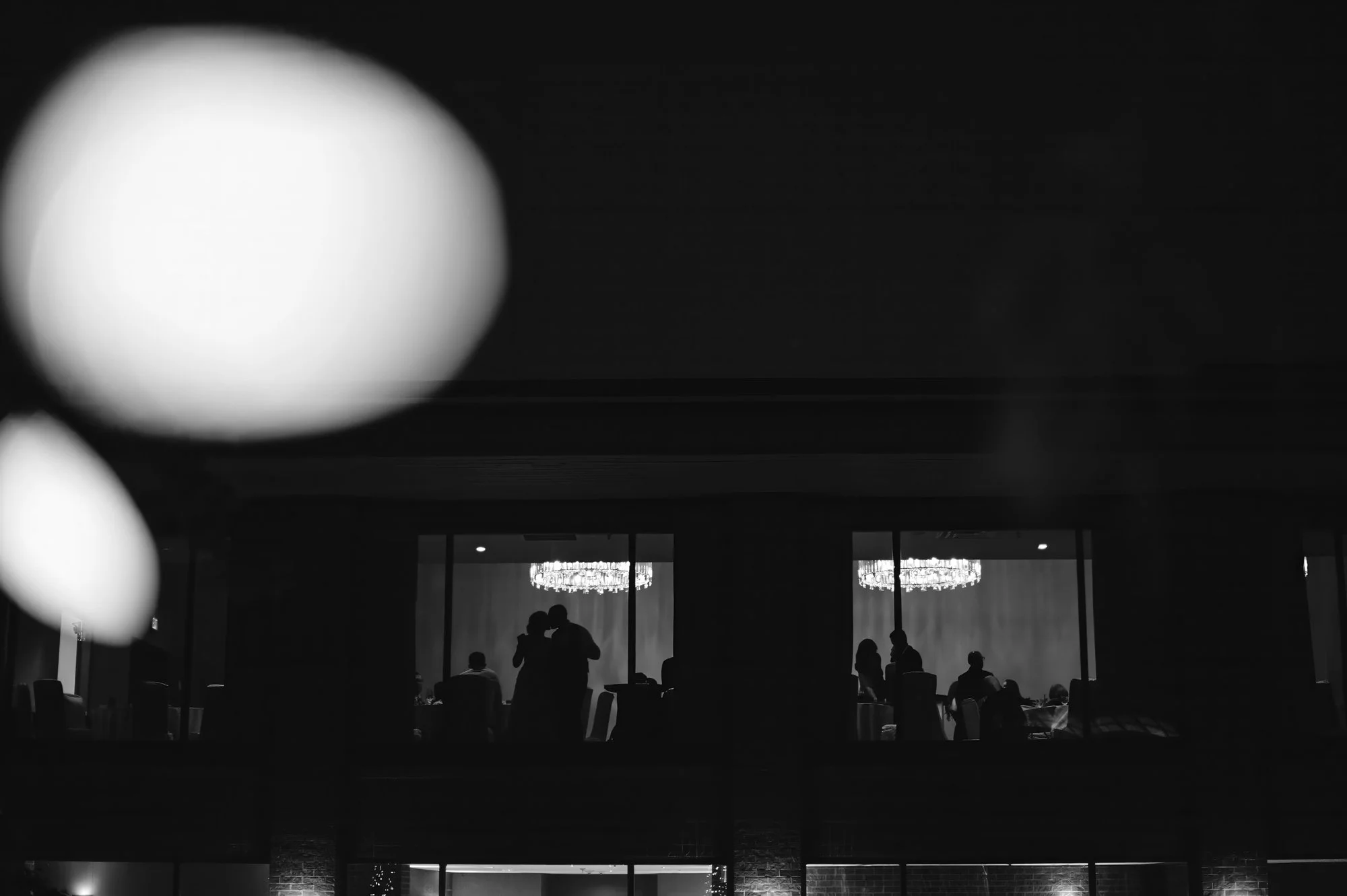Silhouettes of people gathering and talking inside a modern, illuminated venue with large chandeliers, seen through a window or glass wall.