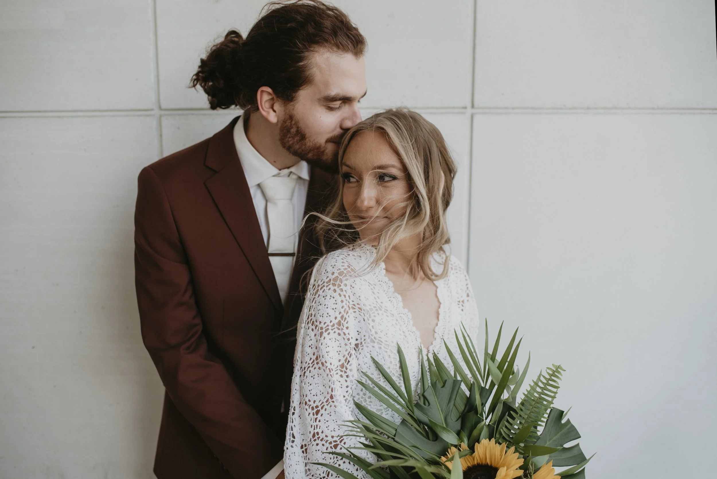 A couple standing close together, the man kissing the woman's temple, woman holding a bouquet of flowers with sunflowers and greenery, both dressed in wedding attire, against a plain wall.