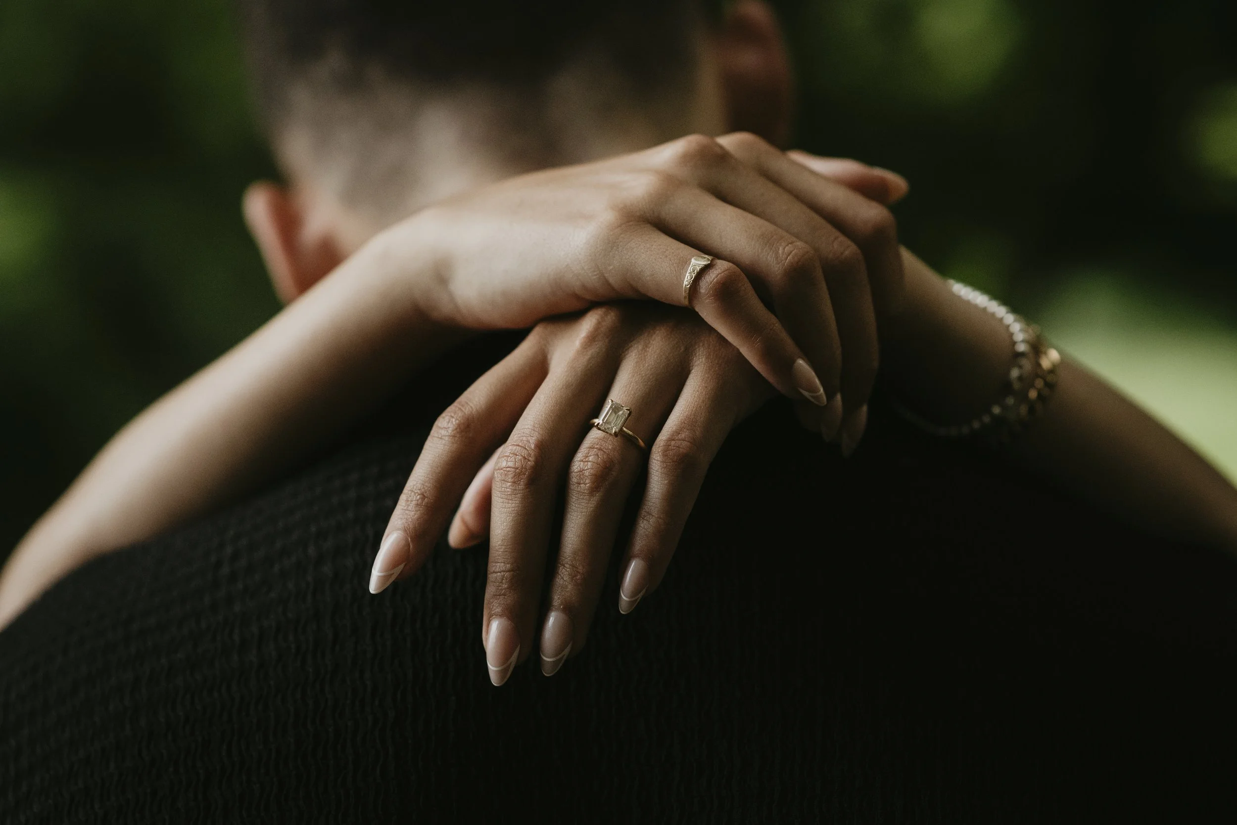 A person with light brown skin and long manicured nails resting their hands on another person's shoulder, showing multiple rings and a bracelet, with a blurred green background.