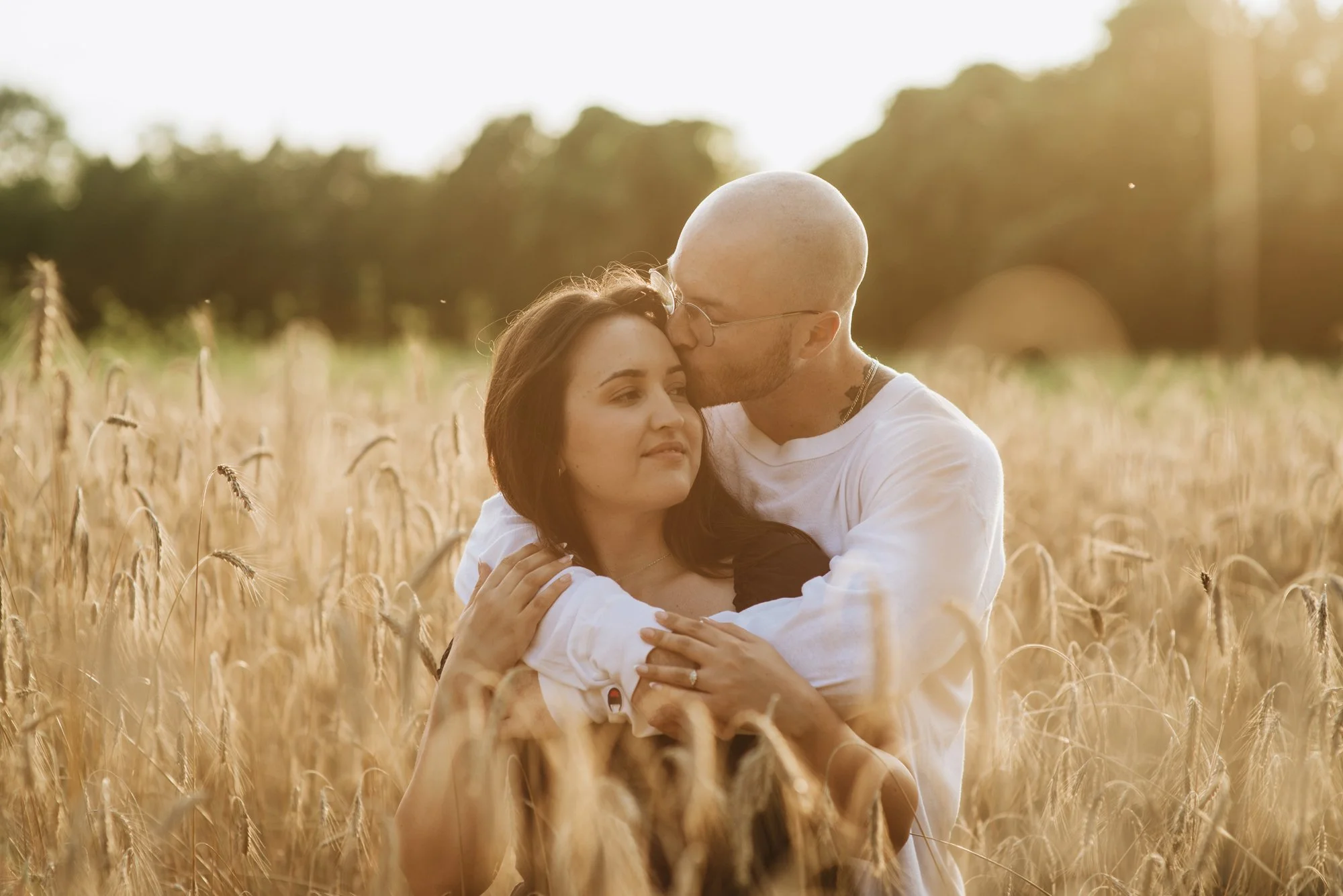 A man with glasses and a bald head kisses a woman on the forehead while embracing her in a wheat field during sunset.