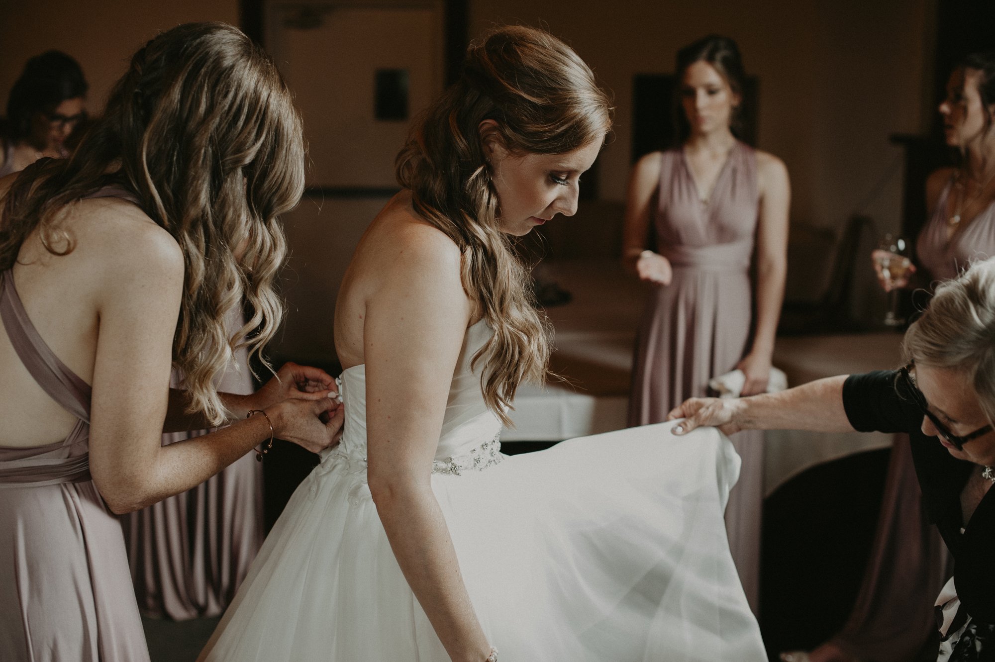 A wedding scene with women in lavender dresses helping a bride in a white wedding gown. The bride is bent over, and her dress is being adjusted by the women around her.