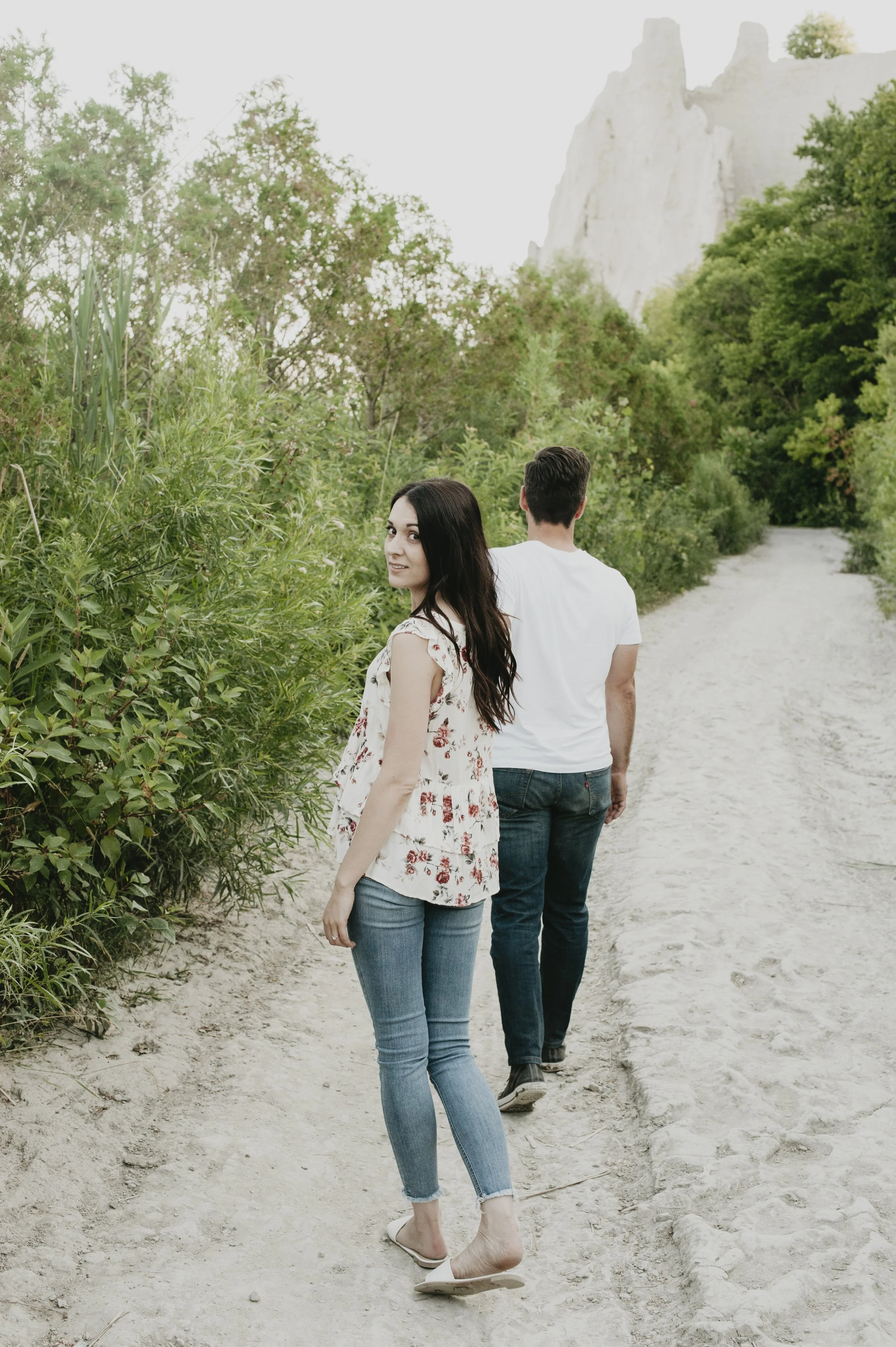 A young woman with long dark hair, wearing a floral blouse, jeans, and white shoes, standing barefoot on a dirt path and looking at the camera. A young man with short dark hair, wearing a white t-shirt and jeans, walking ahead on the same path. She i
