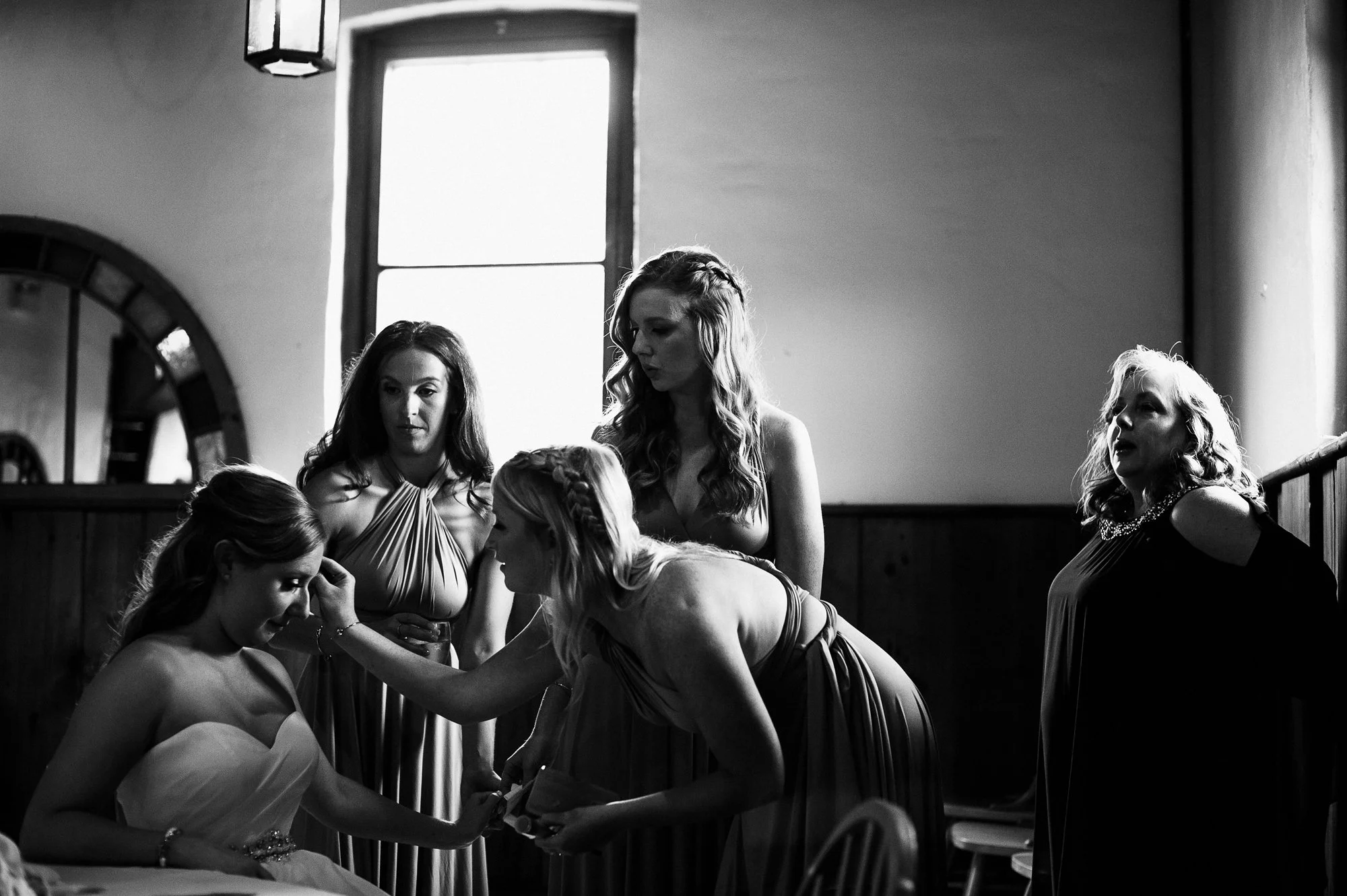Black and white photo of women in formal dresses at a social event, engaging and conversing inside a room with high windows and wooden paneling.