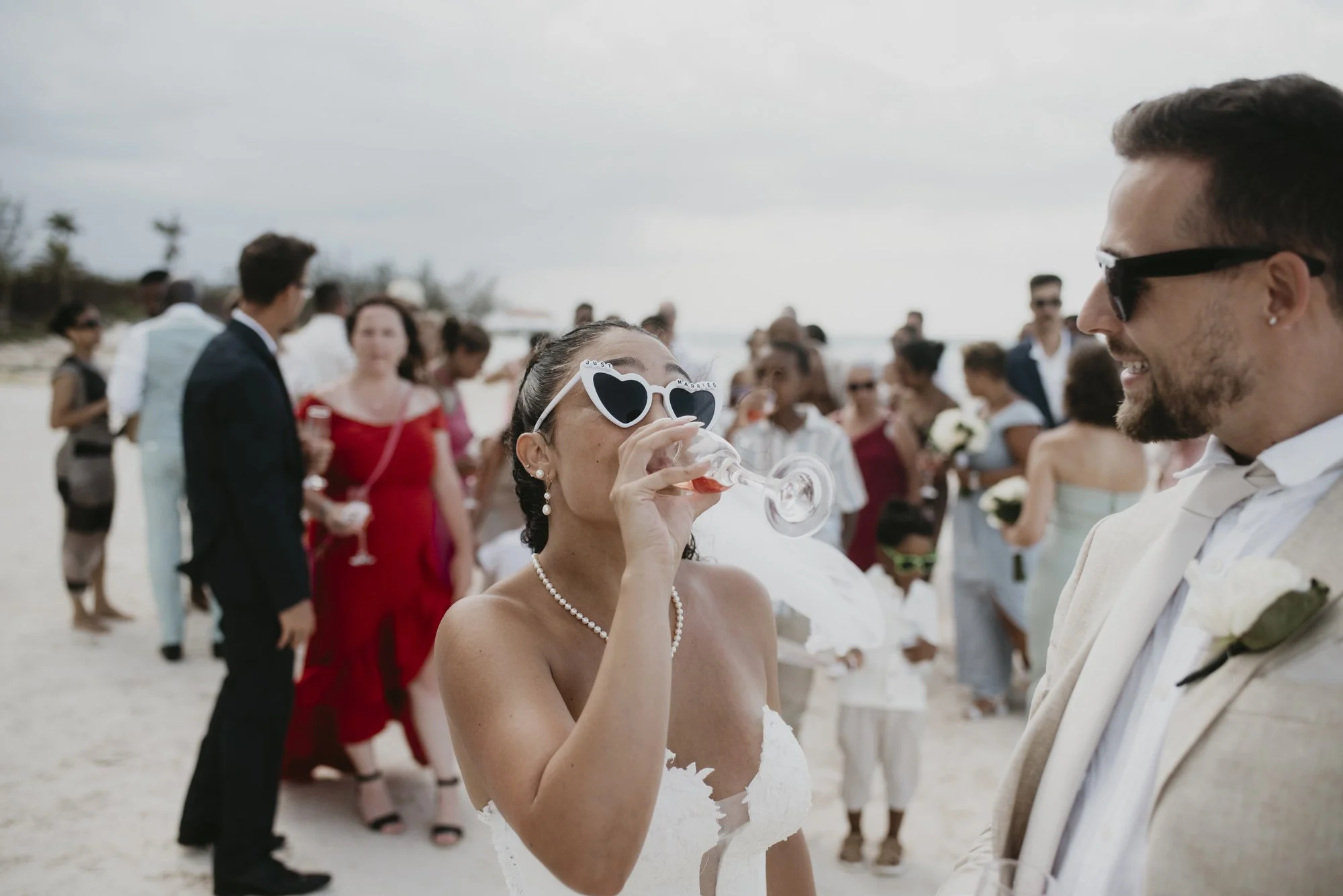 A bride wearing heart-shaped sunglasses on a beach, drinking from a glass, with a smiling groom in a white suit nearby, during her wedding celebration with guests in the background.