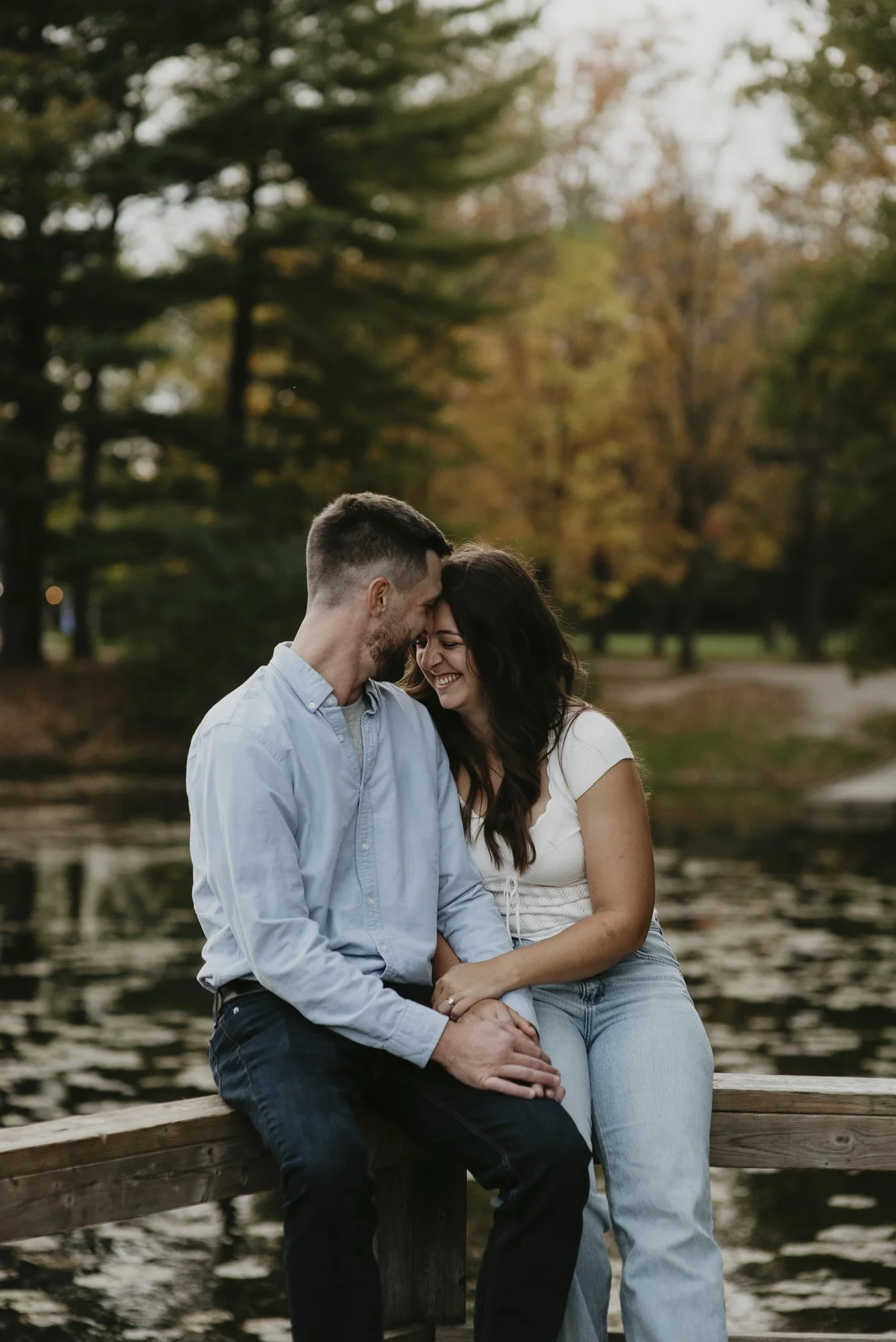 A smiling couple sitting on a wooden dock by a lake, touching foreheads, with trees and fall foliage in the background.