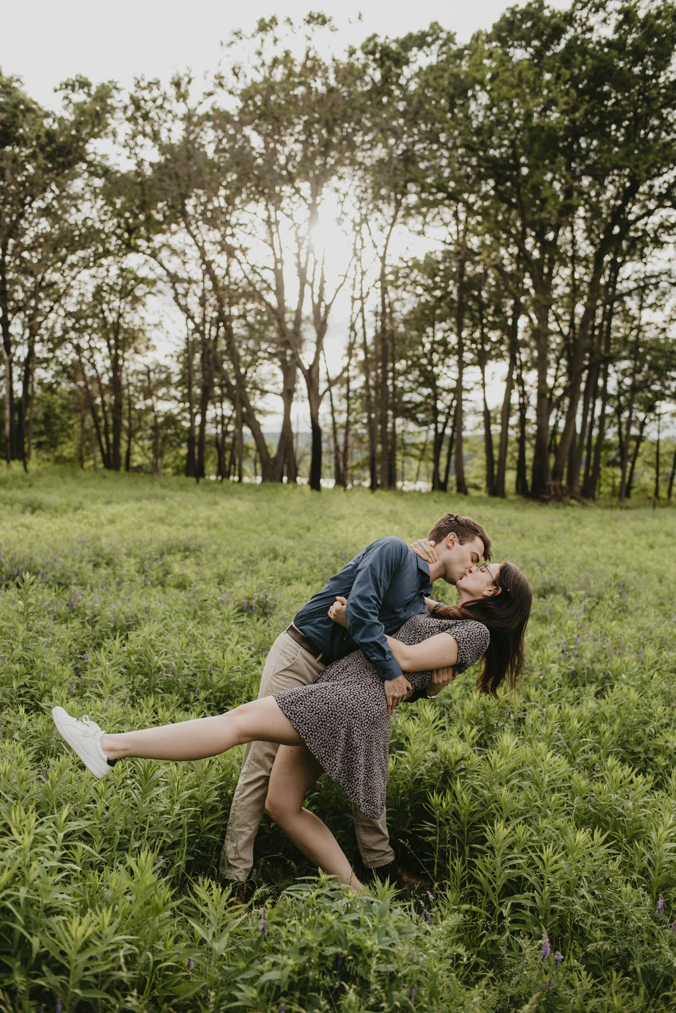 A couple sharing a kiss while one dips the other in a field of greenery during sunset.