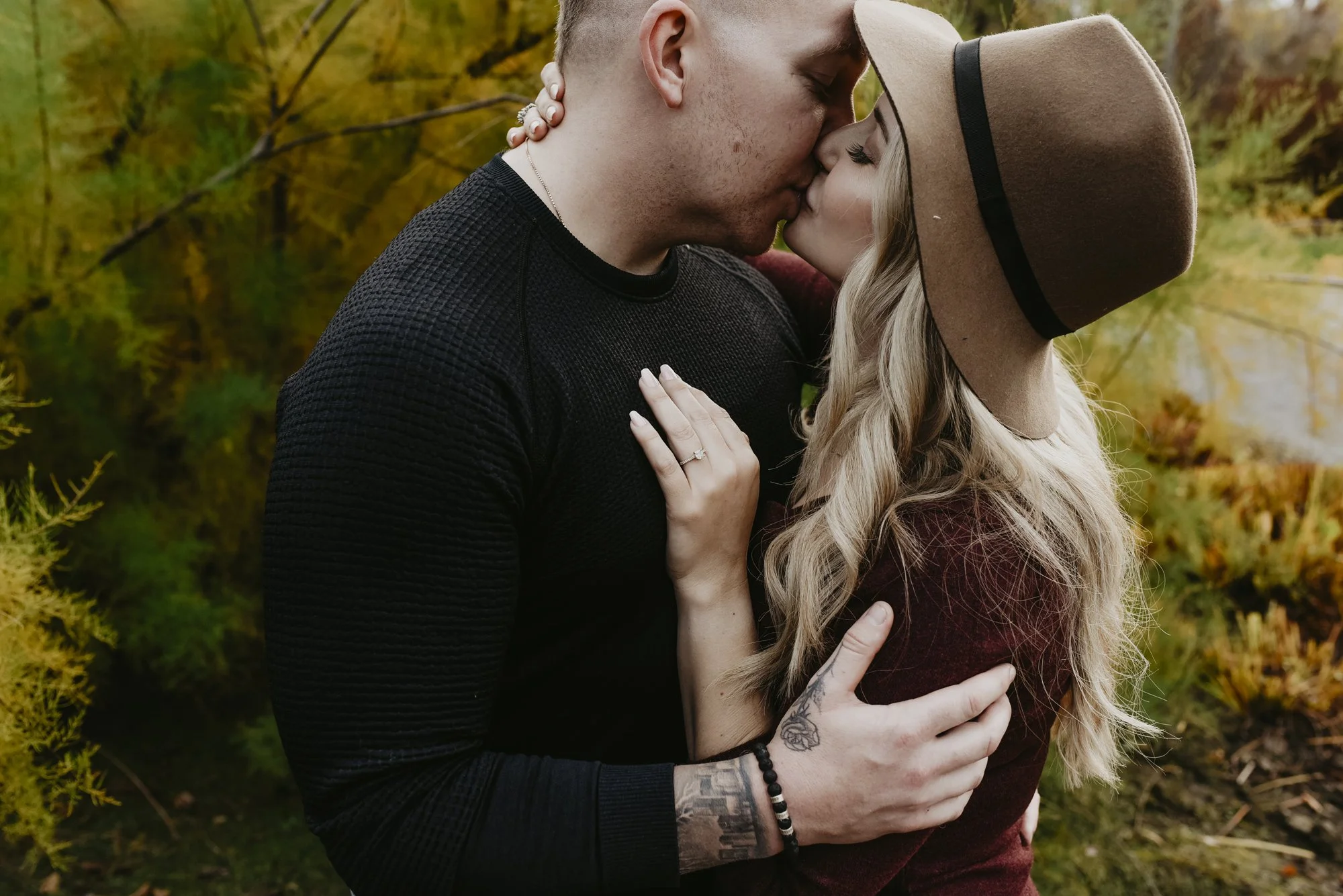 A couple sharing a kiss outdoors during autumn. The man is wearing a black textured long-sleeve shirt, and the woman is wearing a brown hat and a maroon coat. They are embracing amidst fall foliage with yellow and orange leaves.